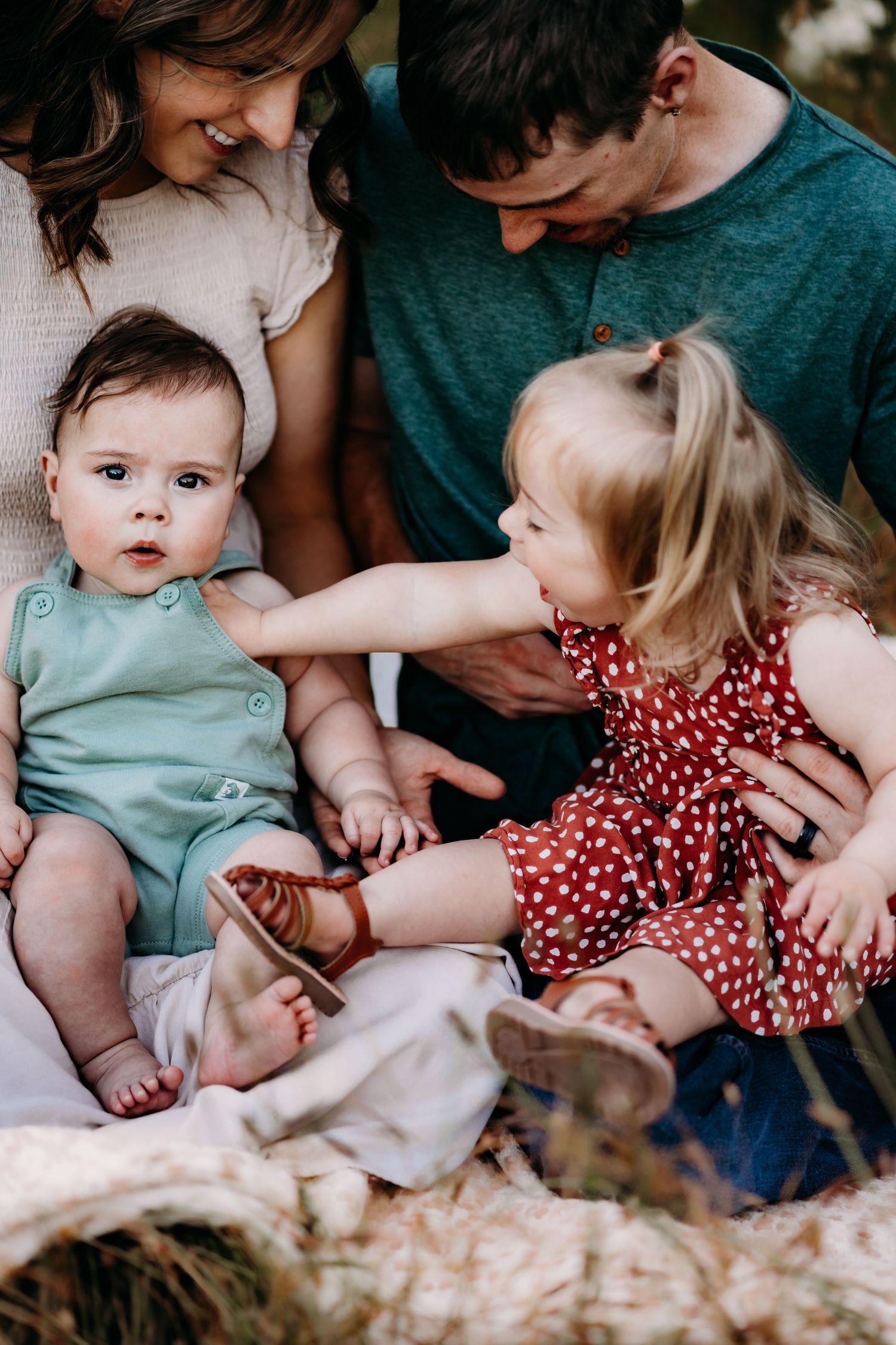 A young family with two toddlers sitting together outdoors, as the older blonde girl in a red polka dot dress reaches for her baby sibling.
