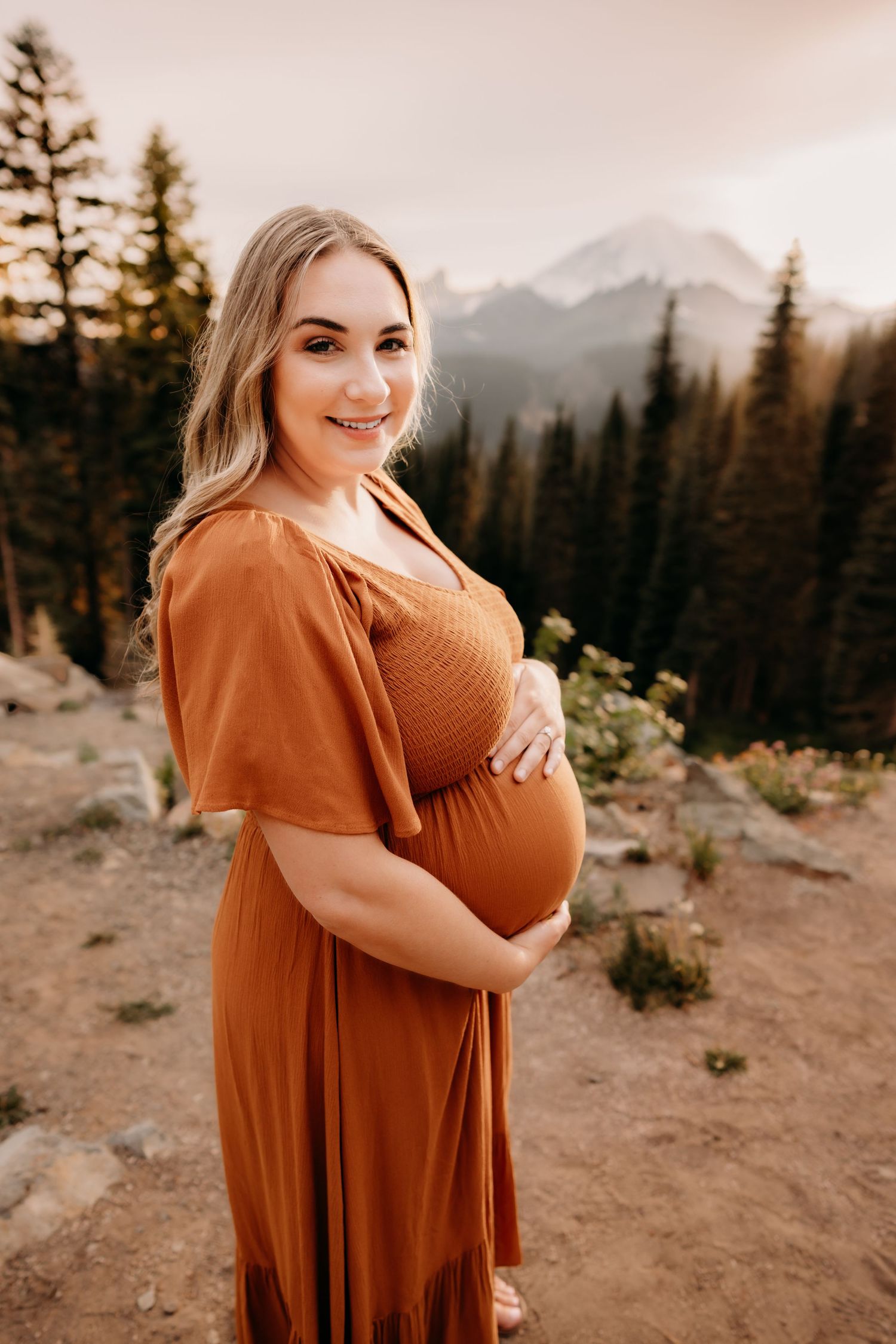 Woman in orange dress stands on a cliff surrounded by tall trees with Mt. Rainier in the background for maternity photos at Tipsoo Lake.