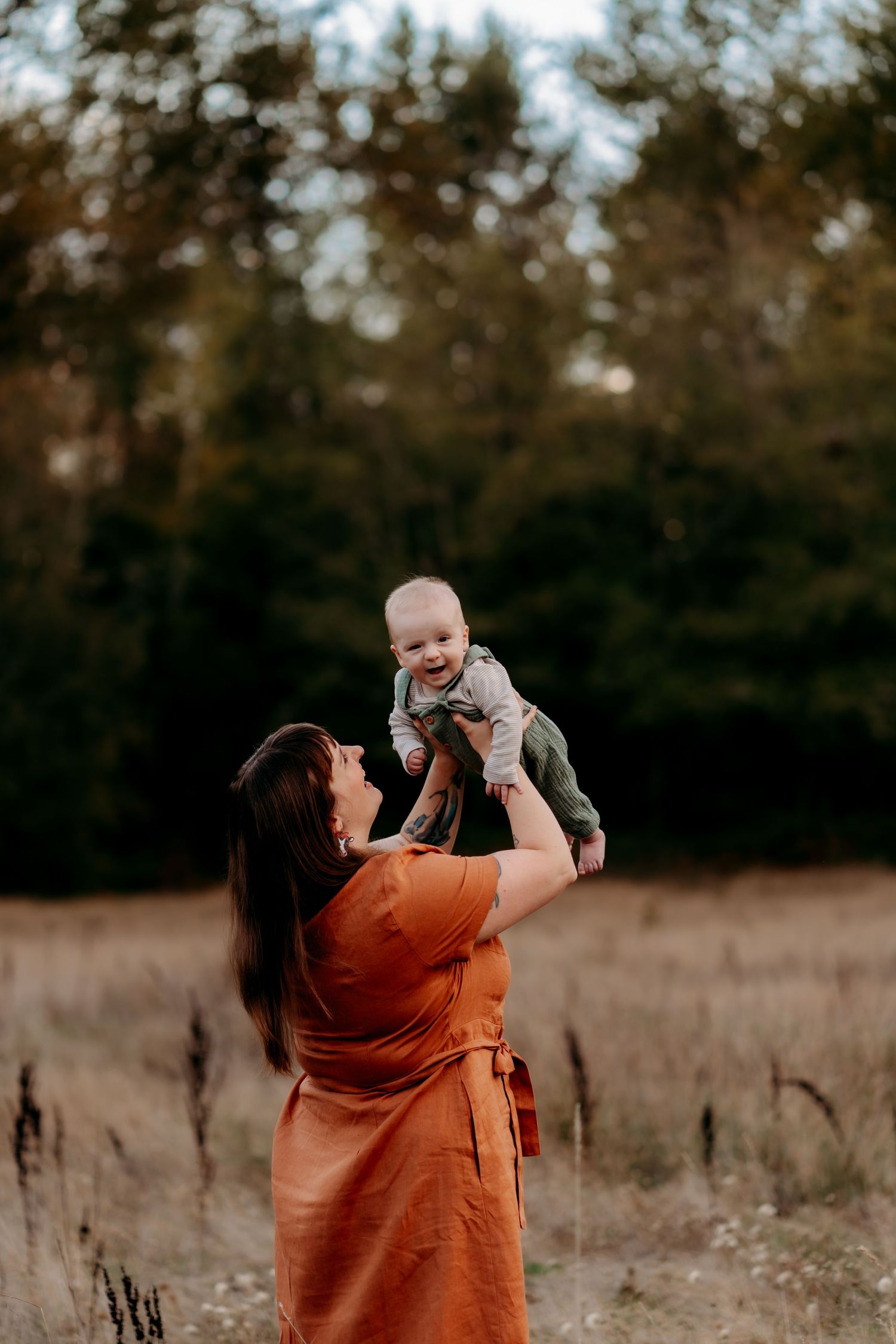 A mother in an orange dress joyfully lifts her smiling baby in a sunlit autumn field with trees in the background during family photos.