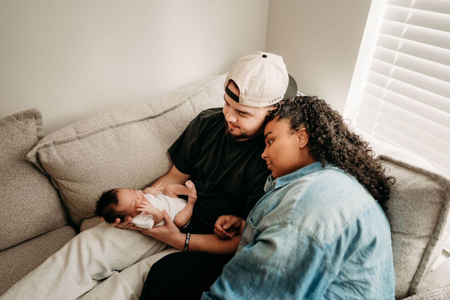 A couple sits on a gray couch sharing a tender moment with their newborn baby in a softly lit room during newborn photo session.