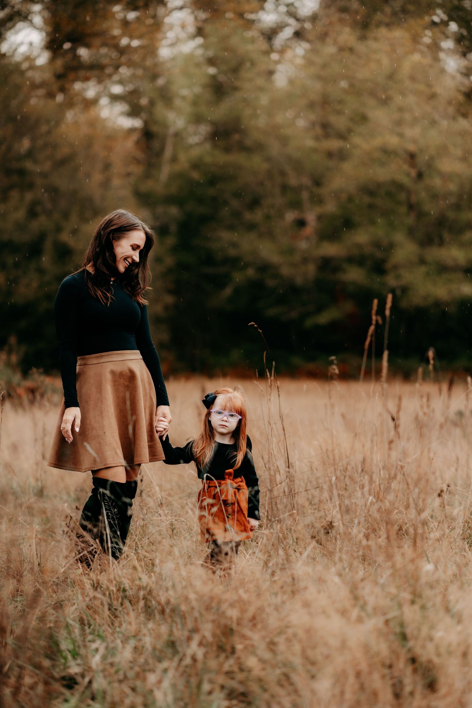 A mother and young daughter hold hands walking through a golden autumn field with forest backdrop at fall family photos in Puyallup, Wa.