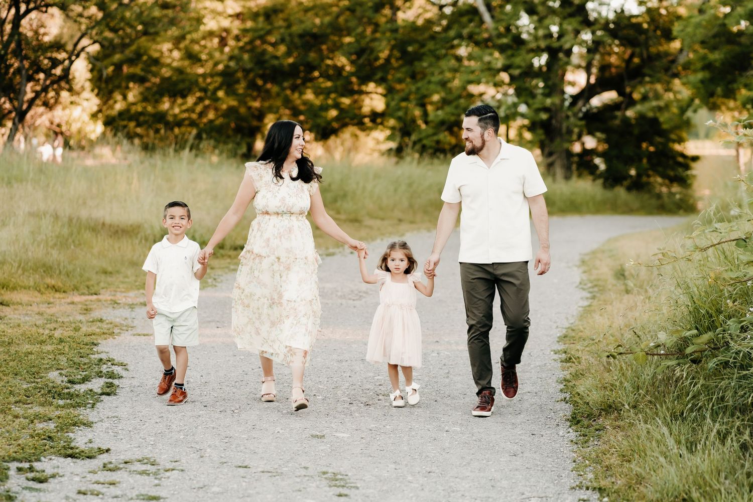 Family of four walking on a gravel path in a sunny park, parents holding hands with young children during family photos in Tacoma, WA.