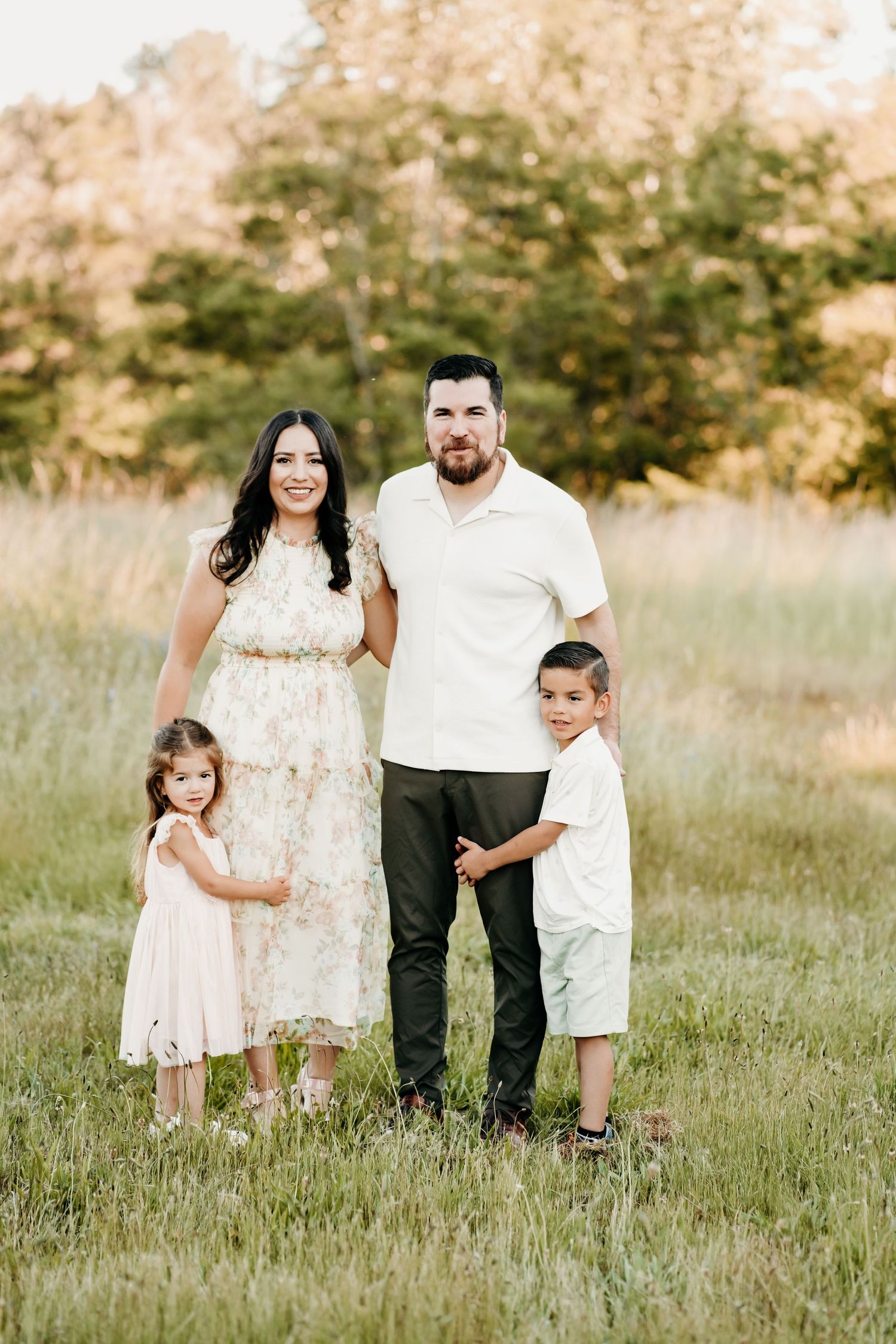 Family of four poses in a sunlit meadow, parents with two young children in coordinating white and pastel outfits for family photos.