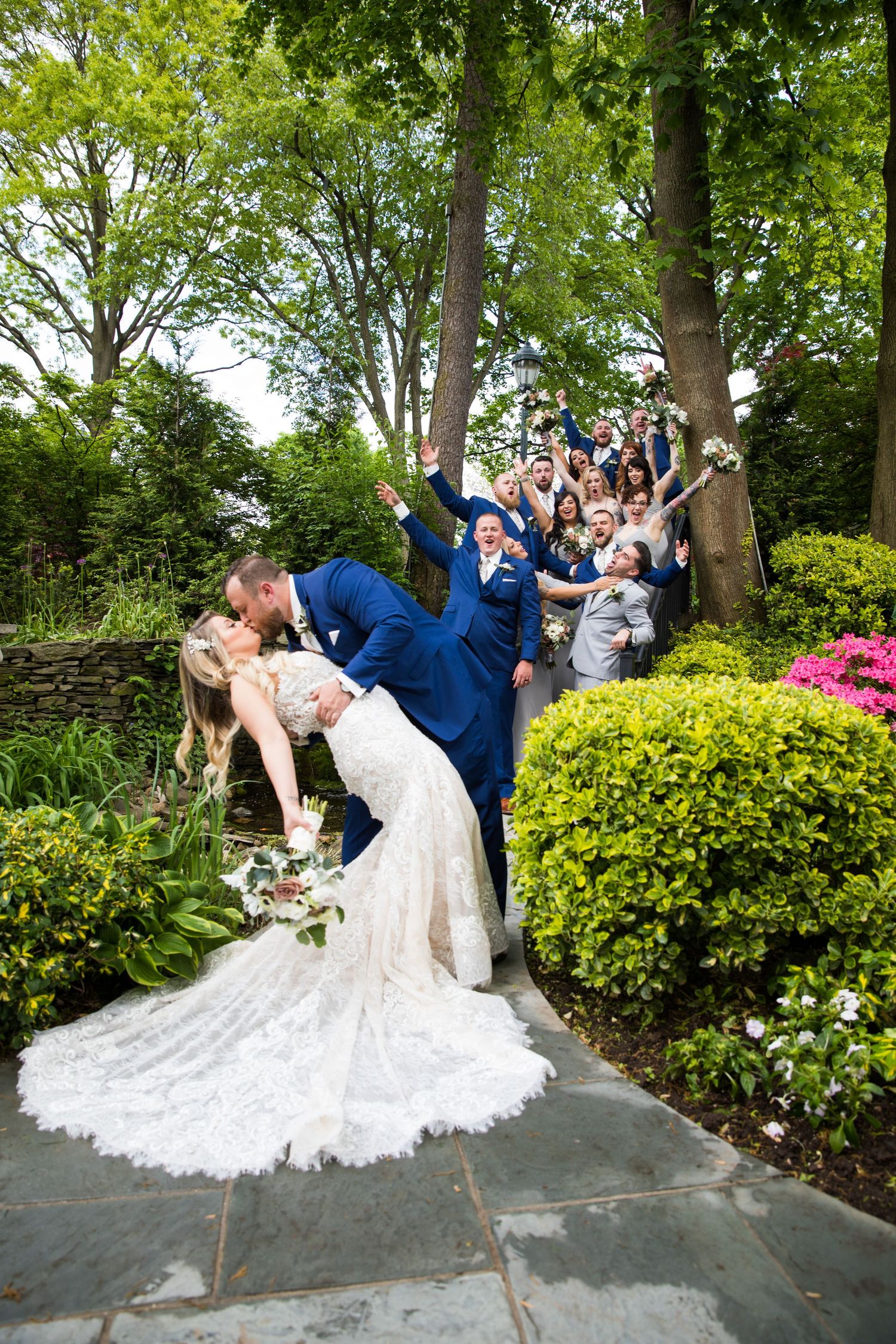 Wedding couple shares dramatic dip kiss in garden setting with guests cheering on stone steps behind them.
