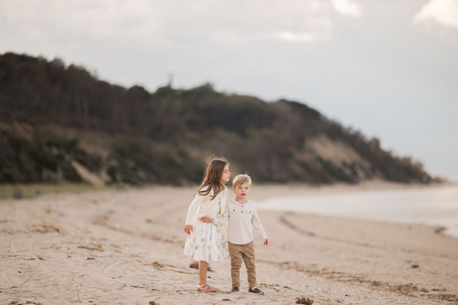 Two children in light colored outfits walk hand in hand along a scenic beach with cliffs.