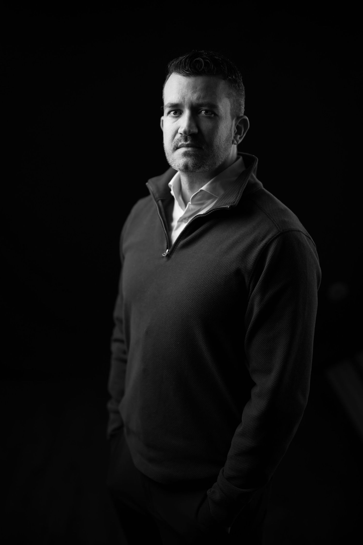Professional black and white portrait of person in dark sweater against black background with dramatic lighting.