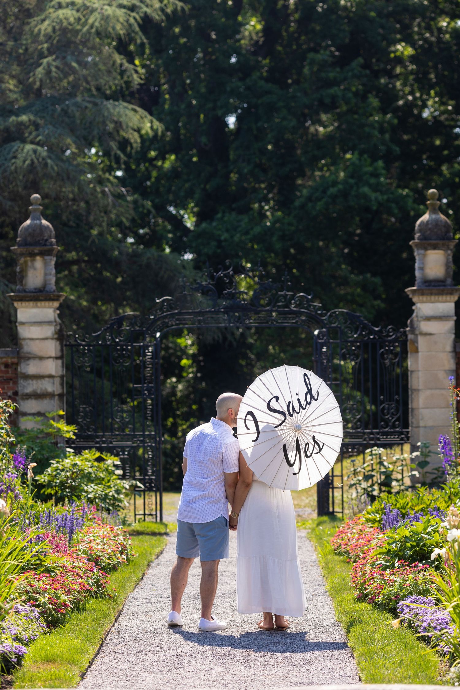 A couple poses with 'I Said Yes' sign on garden path lined with colorful flowers.
