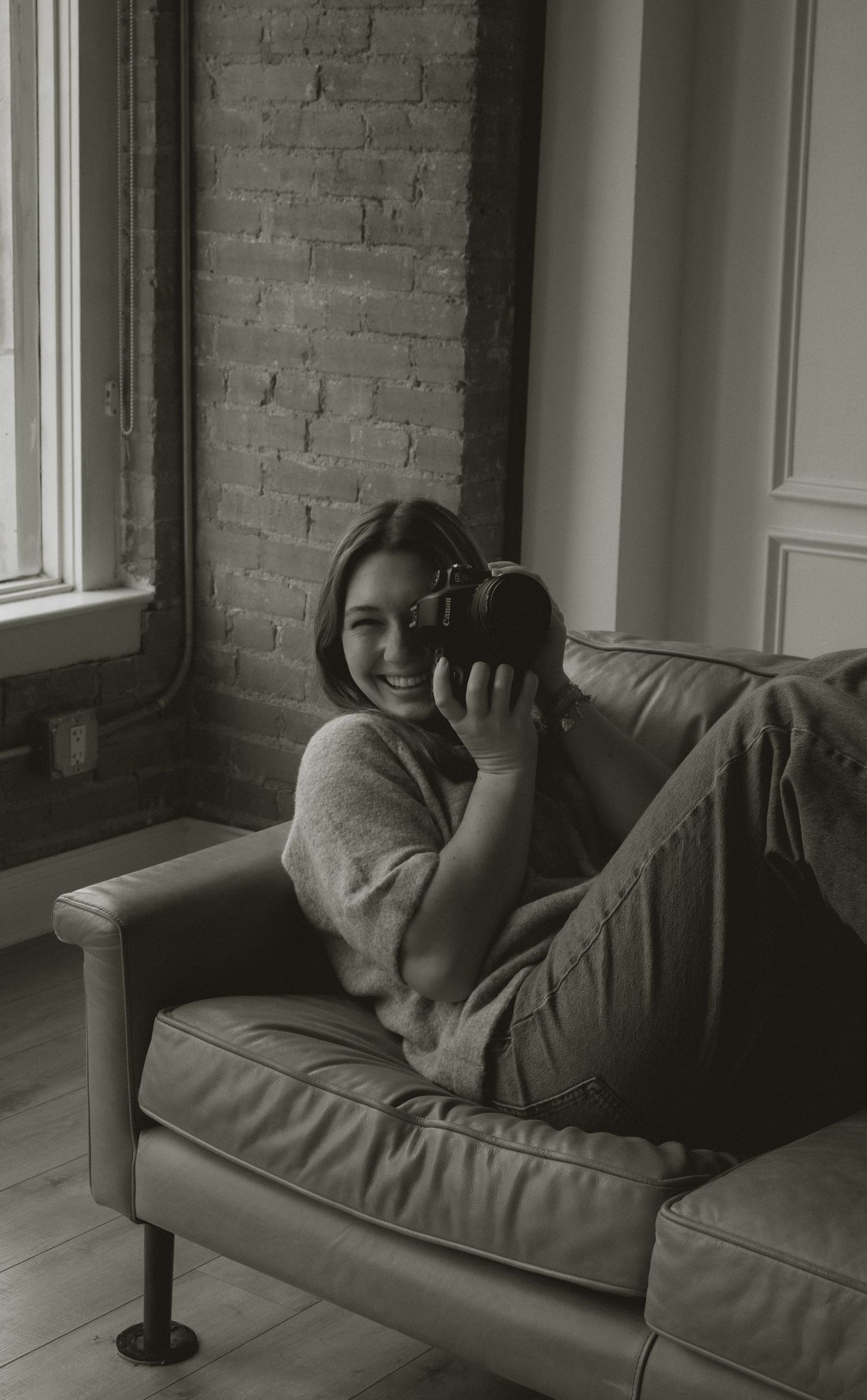 Black and white photo of smiling young woman lying on leather sofa holding a film camera near brick wall.