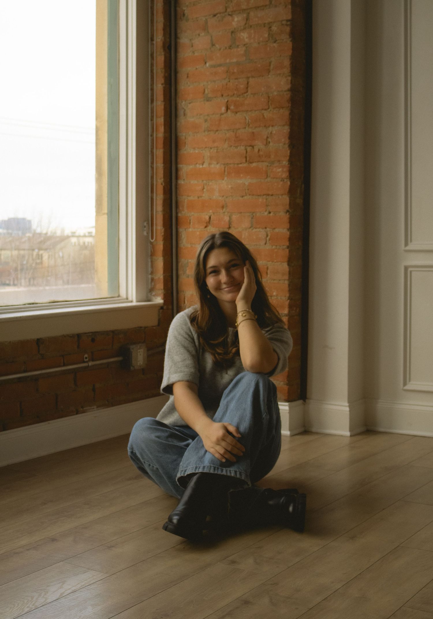 Young woman sitting cross-legged on hardwood floor by large window with exposed brick wall, smiling warmly.
