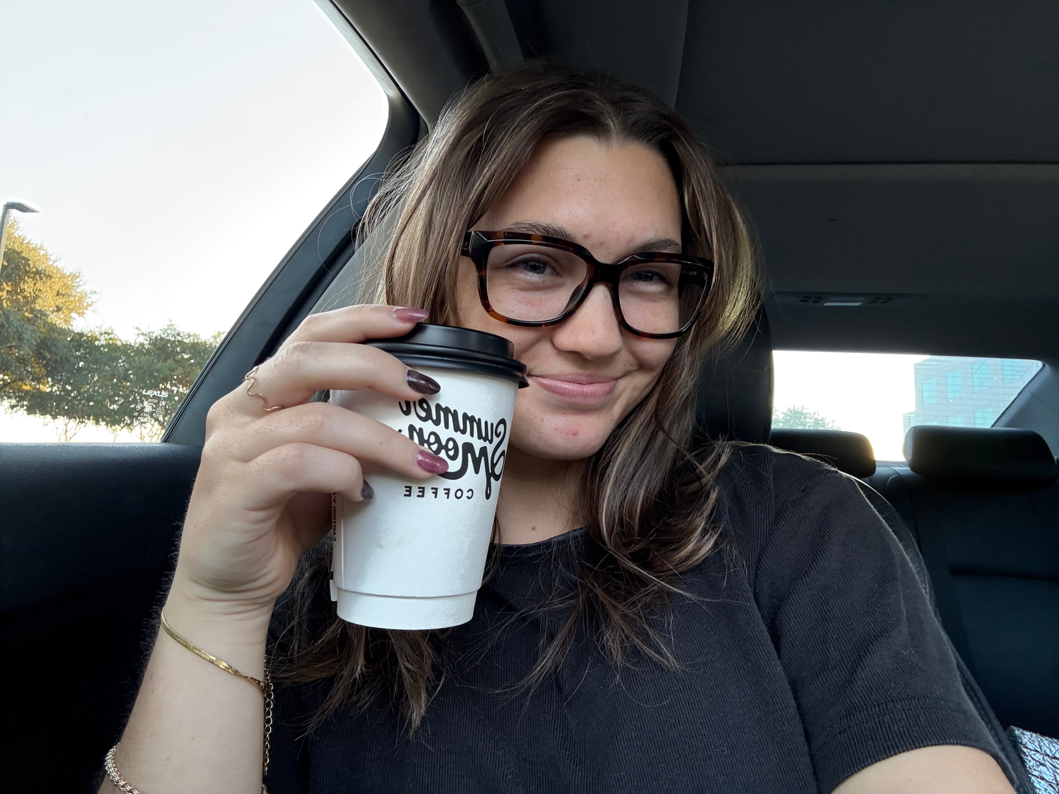 Young woman with glasses smiling and holding a coffee cup inside a car.