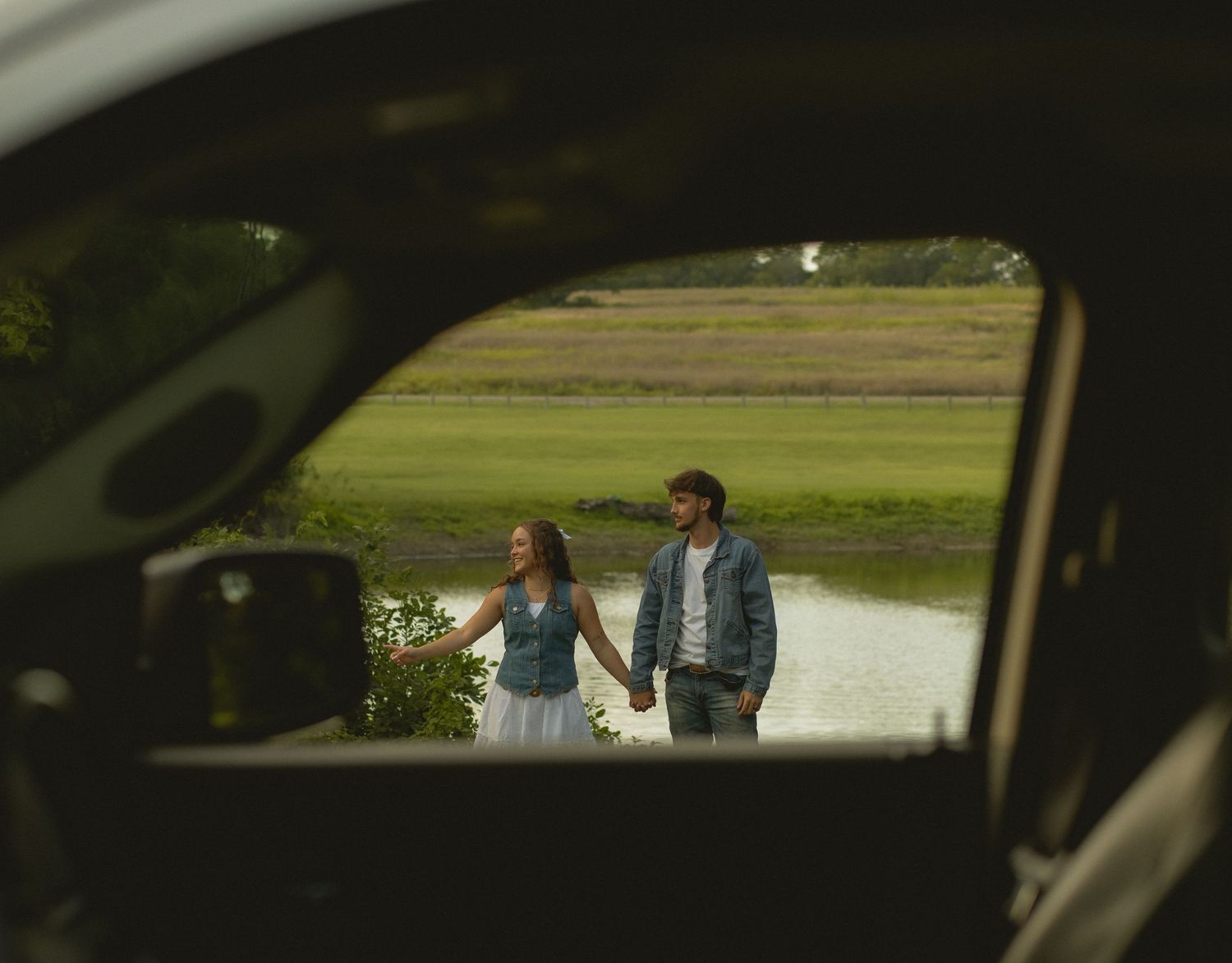 Couple holding hands walking near a field, photographed through a car window frame.