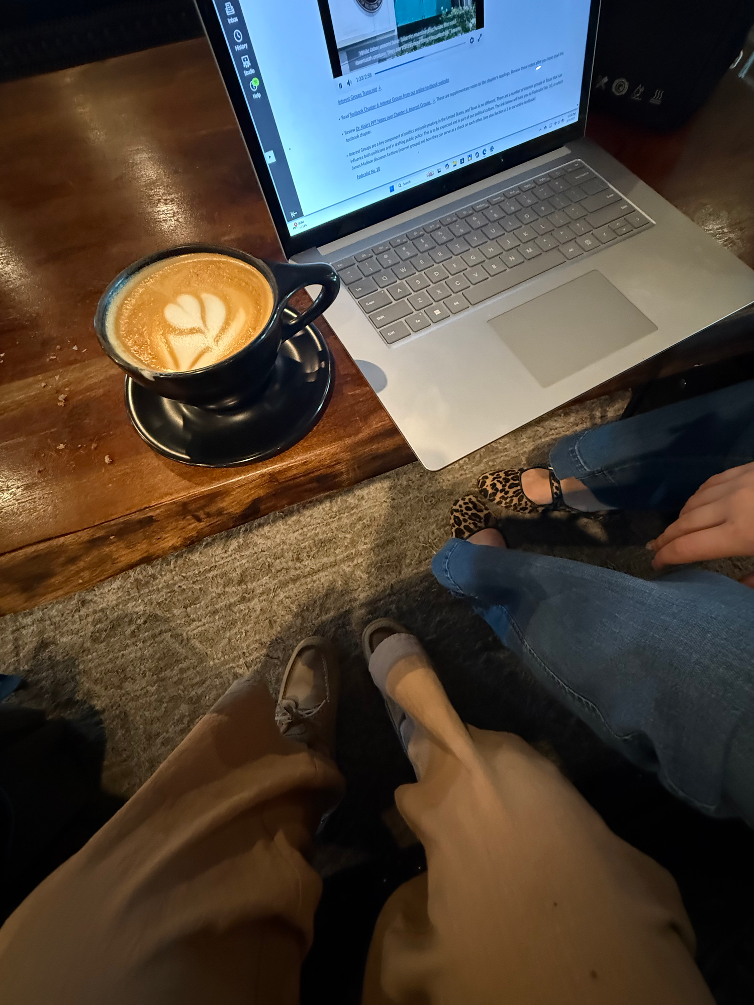 Overhead view of latte art in black mug beside open laptop and two people sitting at cafe table.