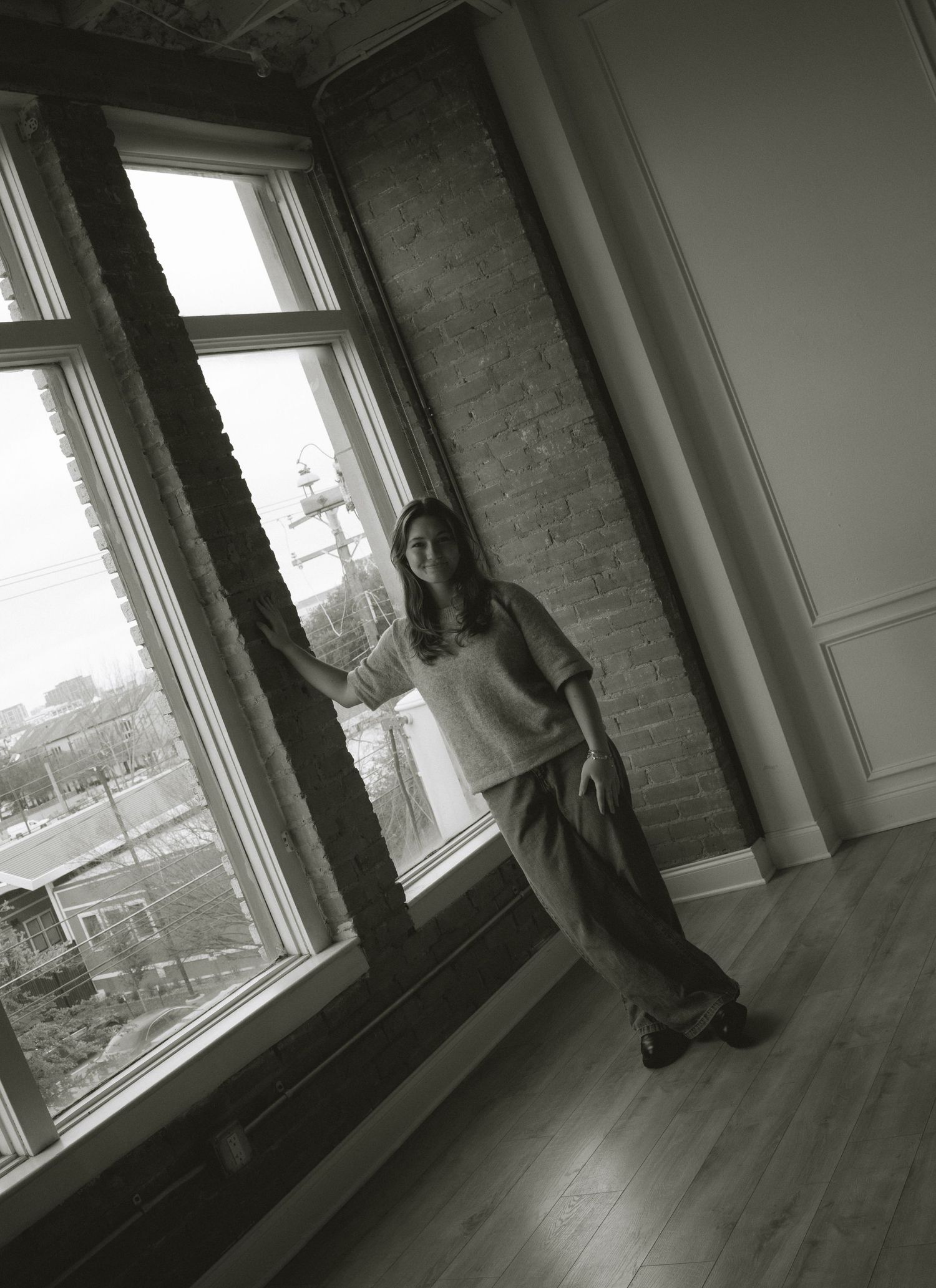 Black and white photo of young woman leaning against brick column by tall windows in minimalist loft space.