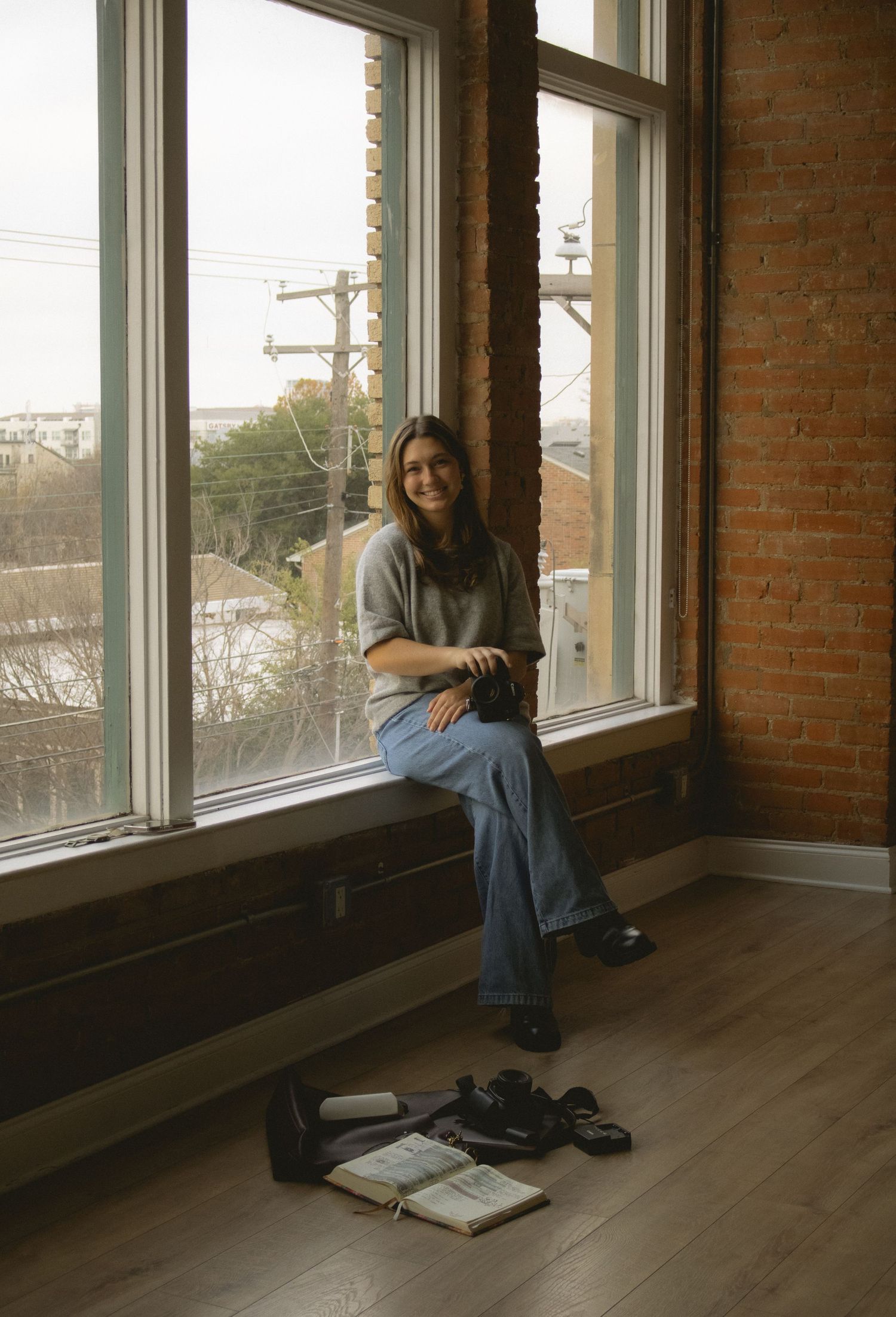 Smiling young woman sitting on window ledge in loft space with exposed brick, camera and bag on floor below.