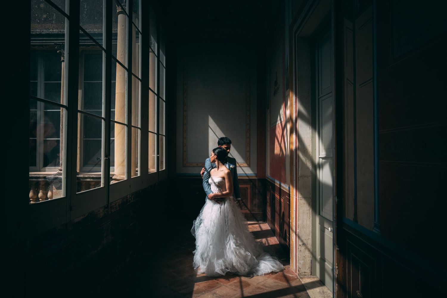 Bride and groom share a romantic embrace in a sunlit hallway with dramatic shadows and large windows.