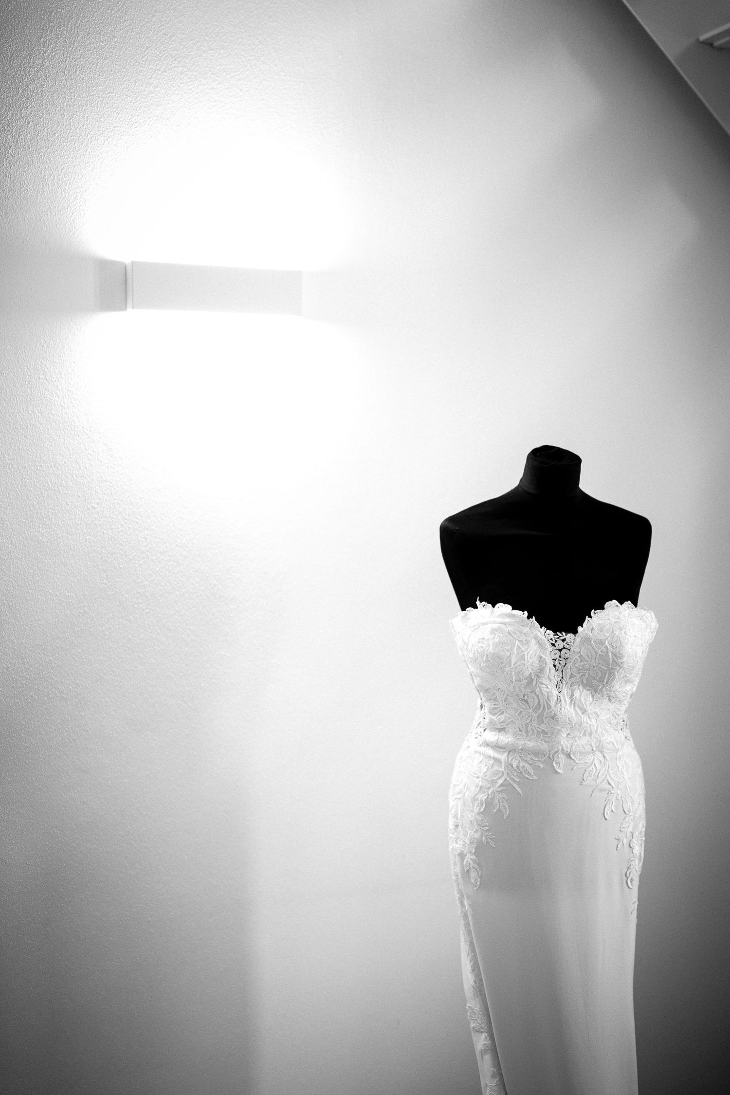 Black and white photo of an elegant strapless lace wedding dress displayed on a mannequin against a white wall.