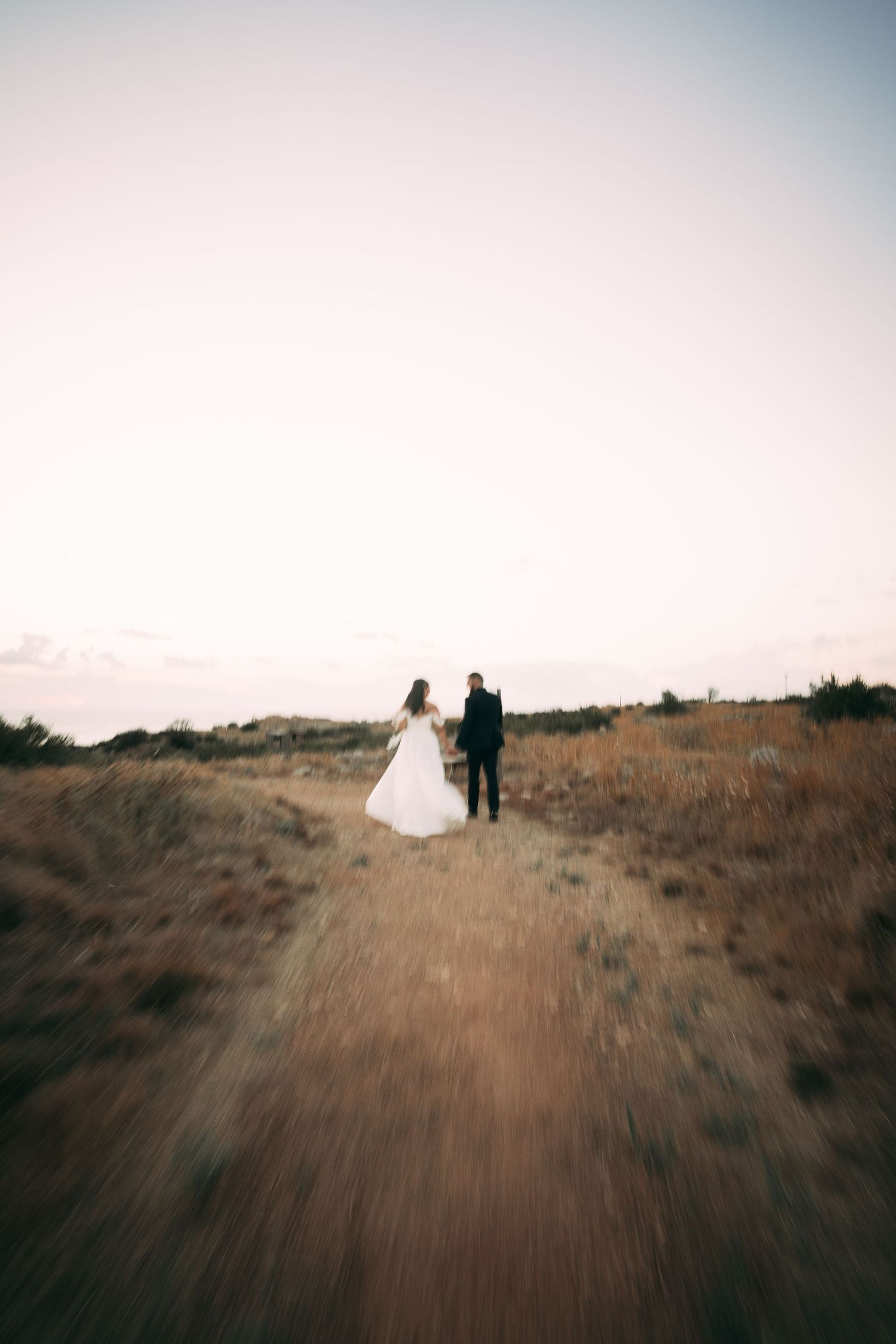 A couple in wedding attire walks along a dirt path on a hill during sunset, captured in a dreamlike motion blur effect.