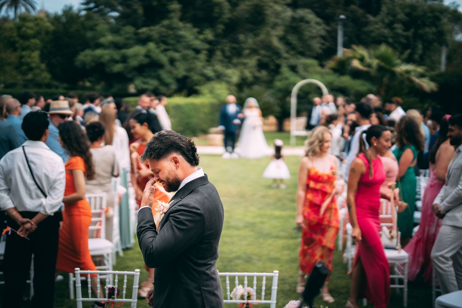 Emotional wedding guest in black suit bowing head during outdoor garden ceremony with guests and couple in background.