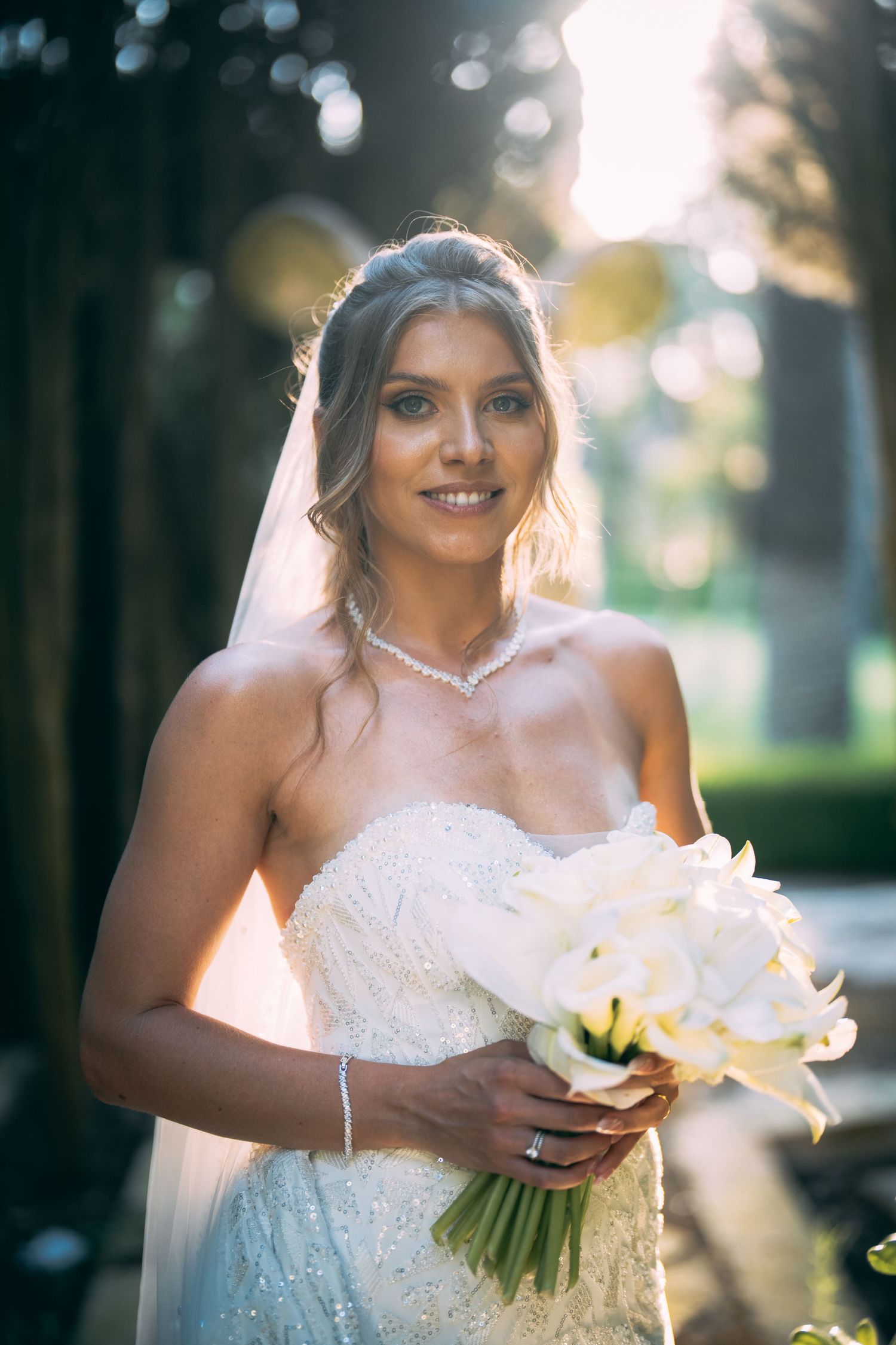 Smiling bride in strapless lace gown holding white calla lily bouquet, wearing veil and pearl necklace outdoors.