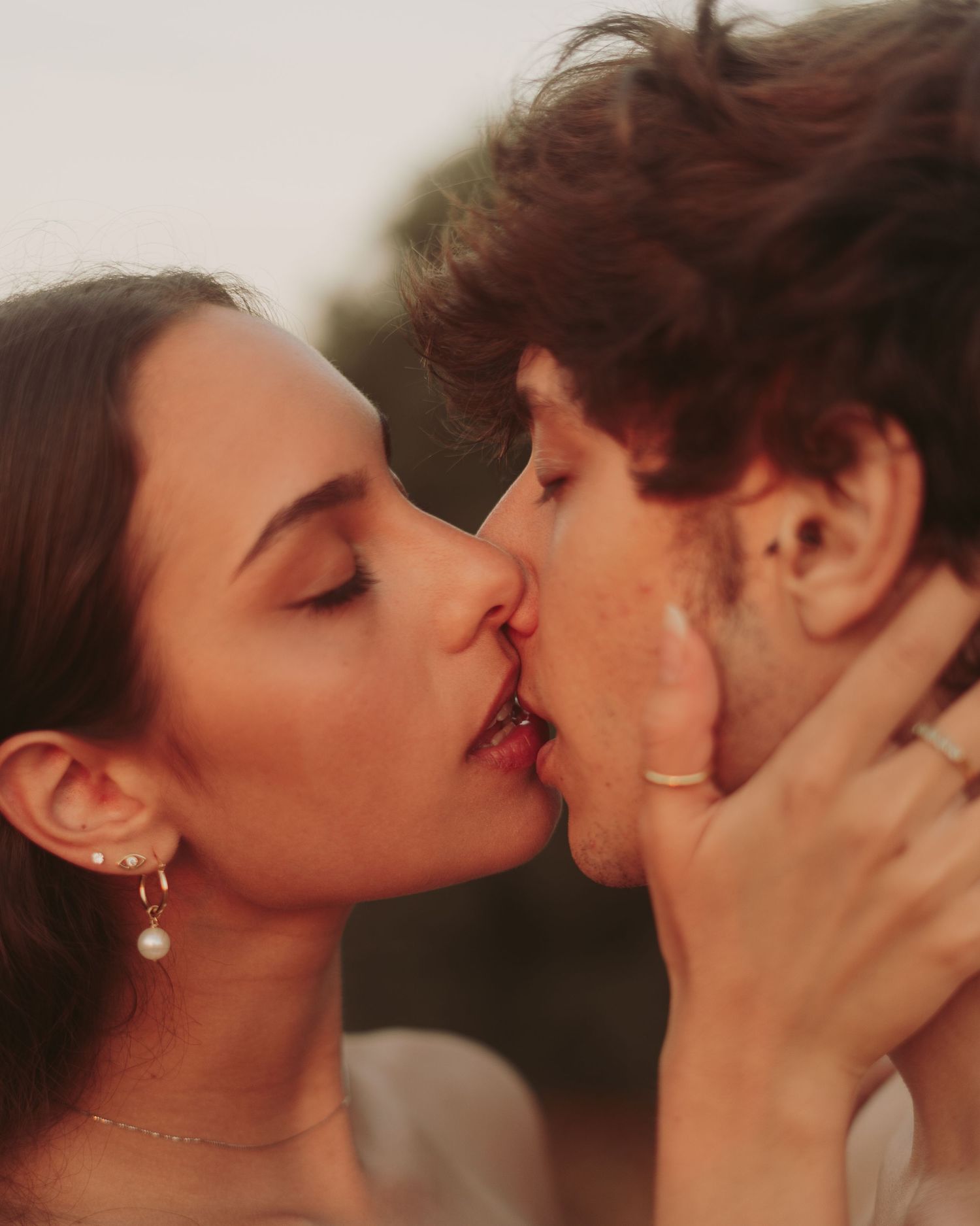 Romantic couple sharing a tender kiss outdoors in warm golden light, woman wearing gold earrings and ring.