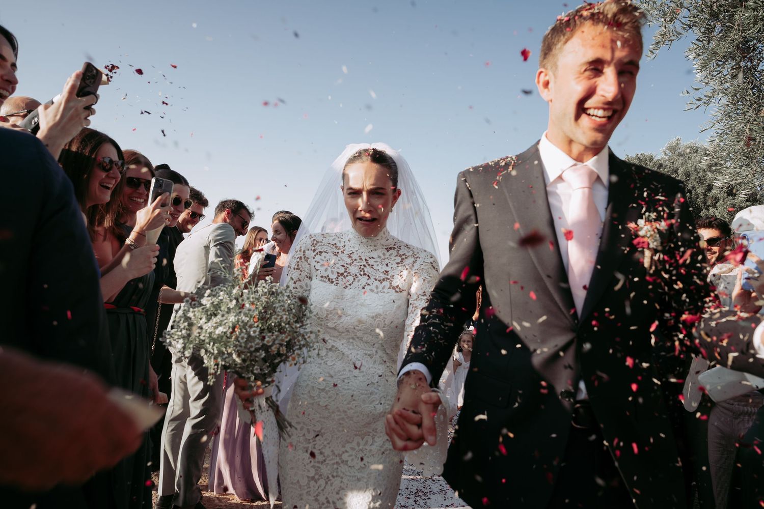 Joyful newlyweds walk through confetti shower after wedding ceremony, bride holding white bouquet.