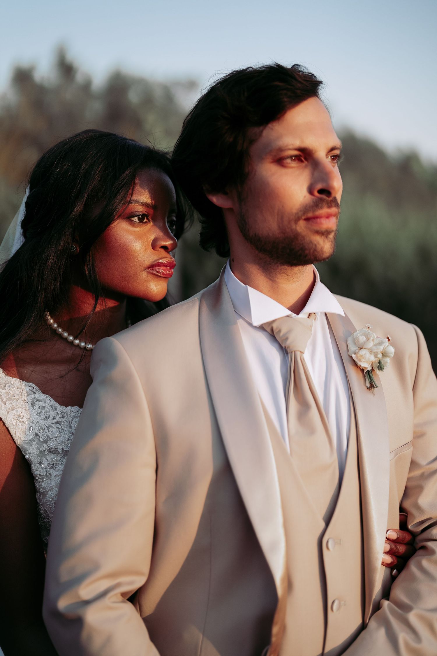 Interracial couple in wedding attire, groom in beige suit with boutonniere, bride in white dress with pearl necklace and veil.