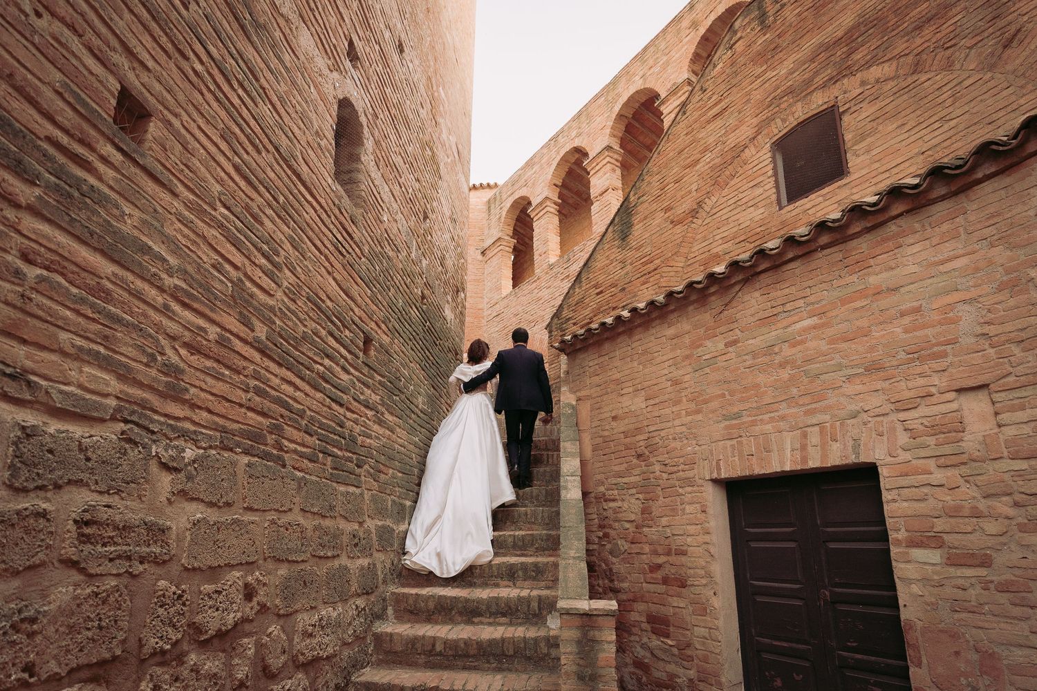 Bride and groom walking up ancient stone steps between brick walls of a historic building.