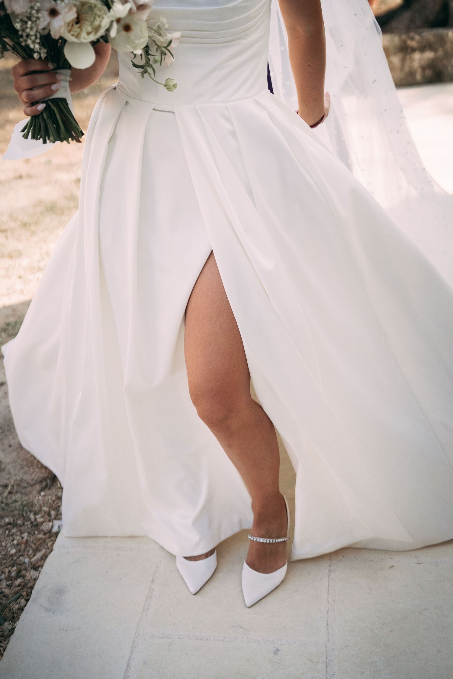 Bride in white slit gown with crystal ankle strap heels holding floral bouquet on stone steps.