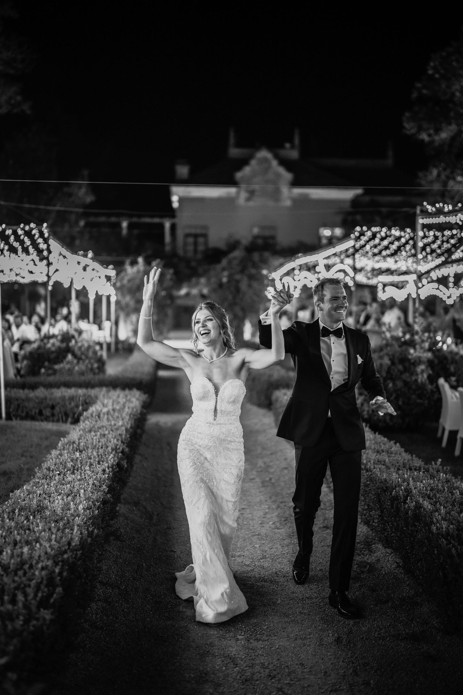 Black and white photo of a joyful bride and groom walking through a garden at night with festive lights.