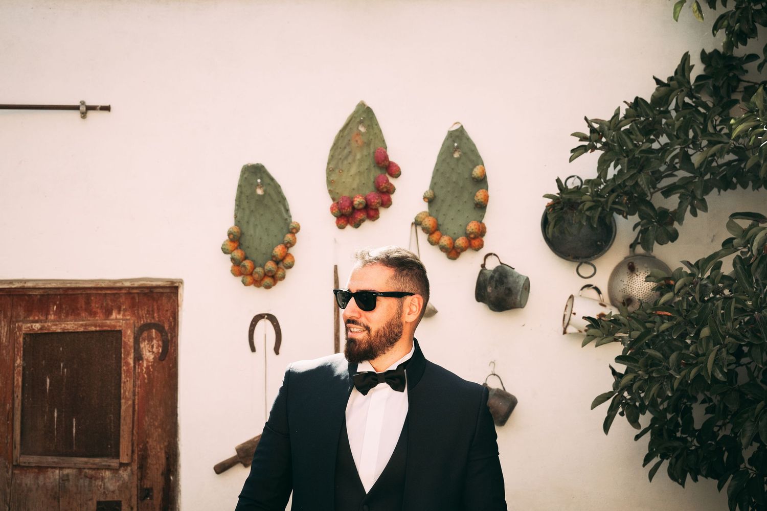 Stylish bearded man in black tuxedo and sunglasses posing outdoors near rustic wall decor and greenery.