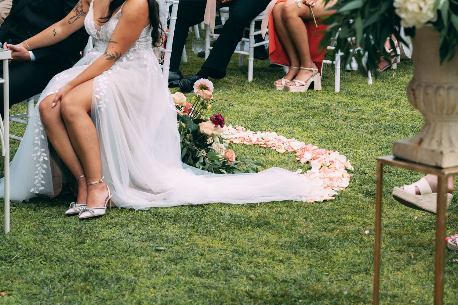 Bride in white gown seated outdoors with rose petal heart shape on green grass at wedding ceremony.