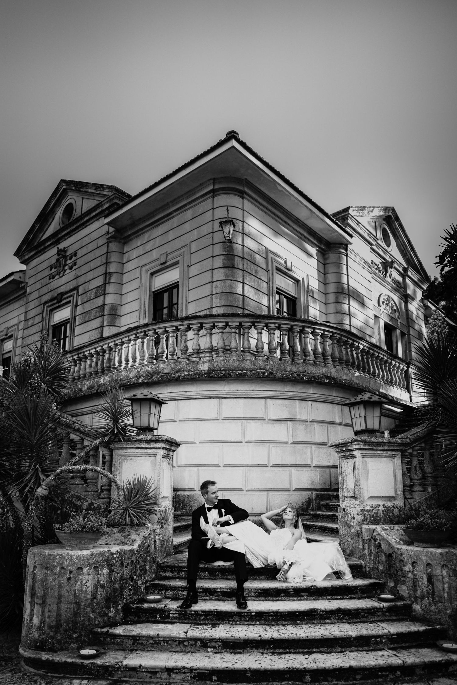 Black and white photo of bride and groom posing on stone steps of an elegant historic mansion.