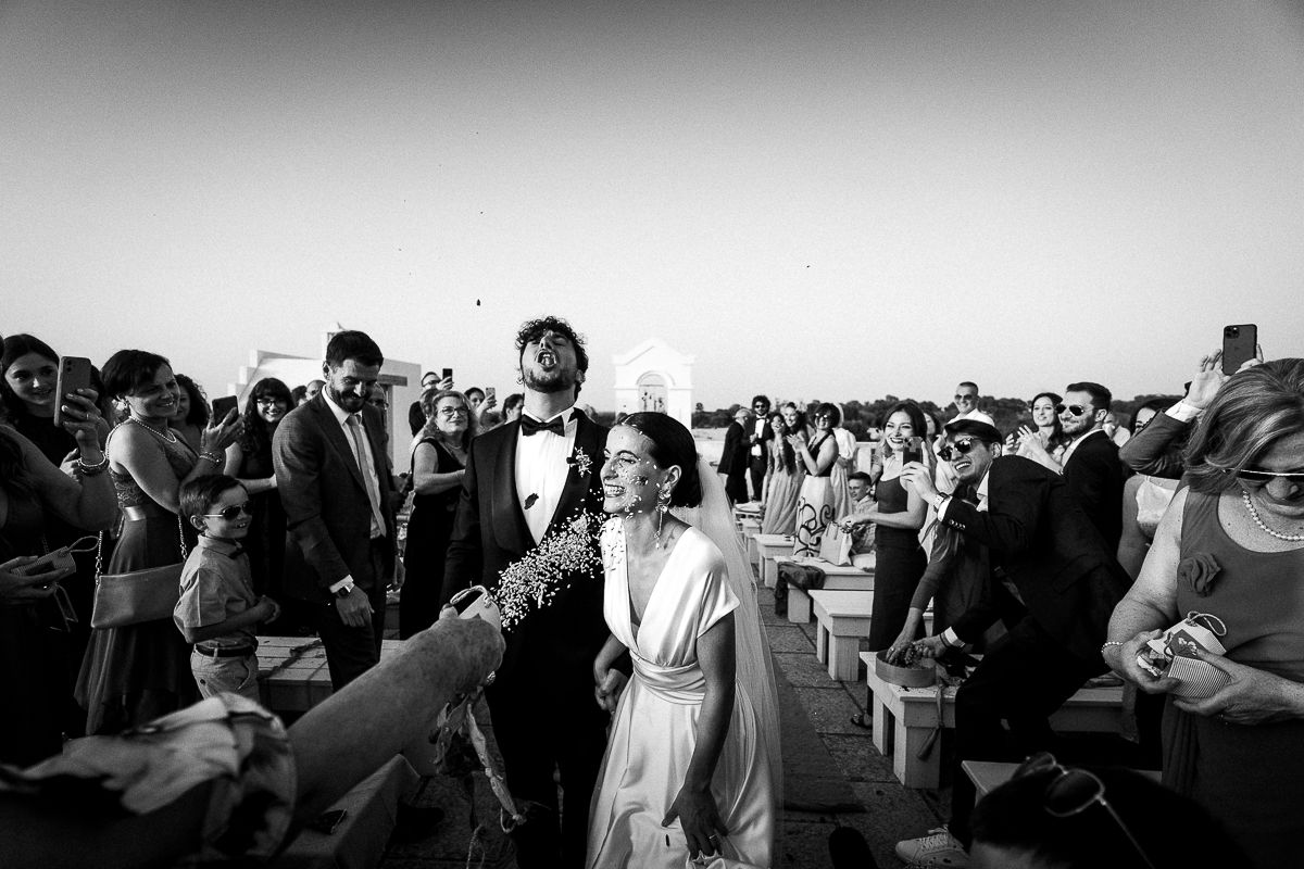 Black and white wedding photo of couple walking through cheering guests outdoors.