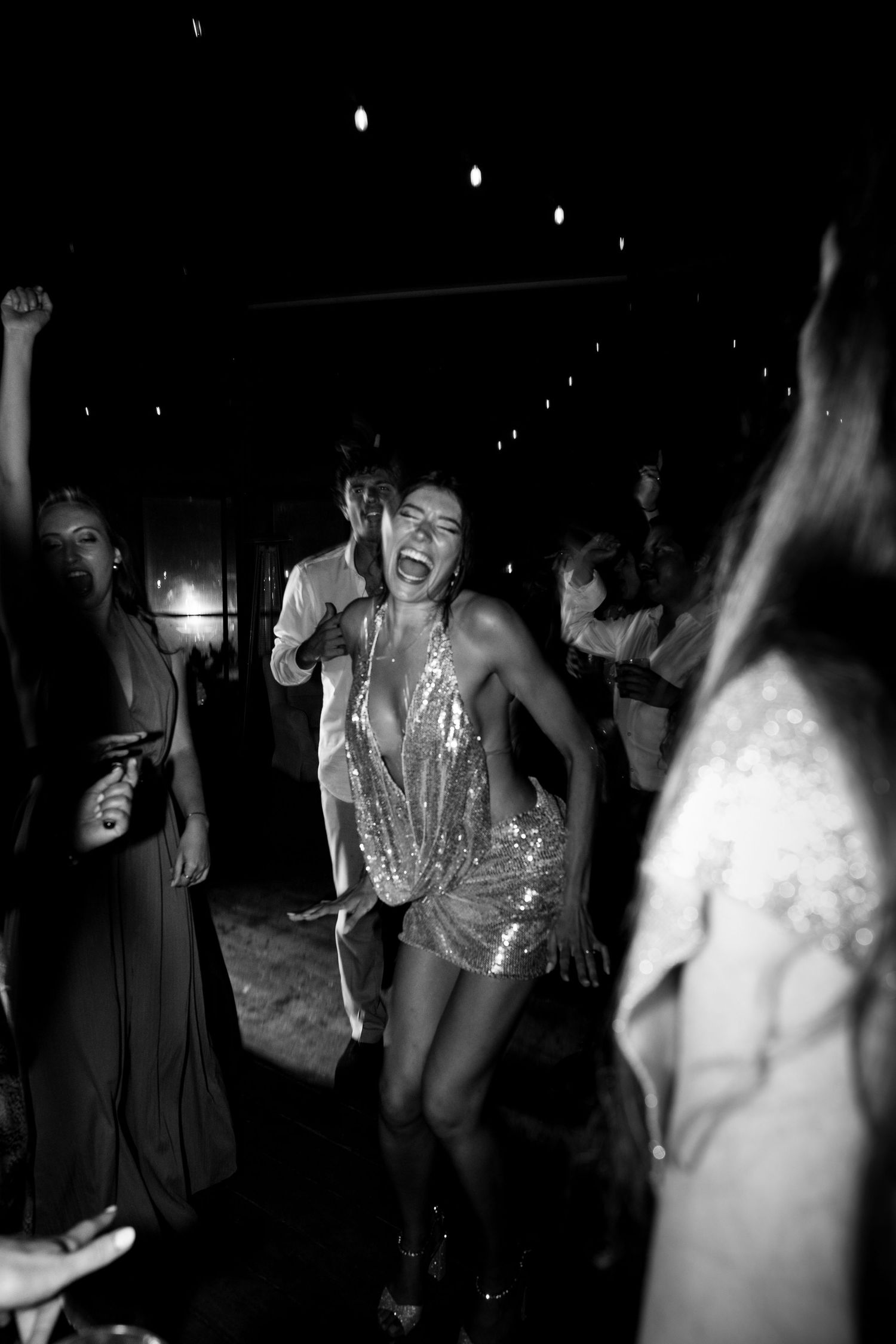 Black and white photo of a woman in a sequin mini dress dancing joyfully at a lively party or nightclub event.