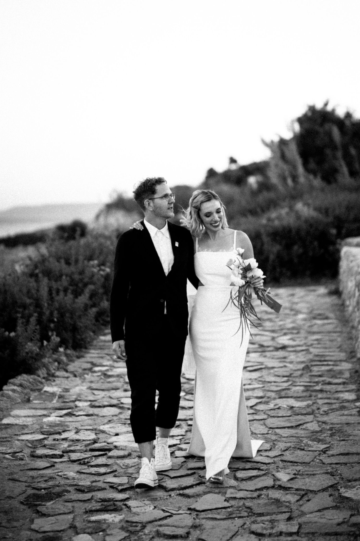 Black and white photo of a bride and groom walking on a cobblestone path, bride holding a bouquet.
