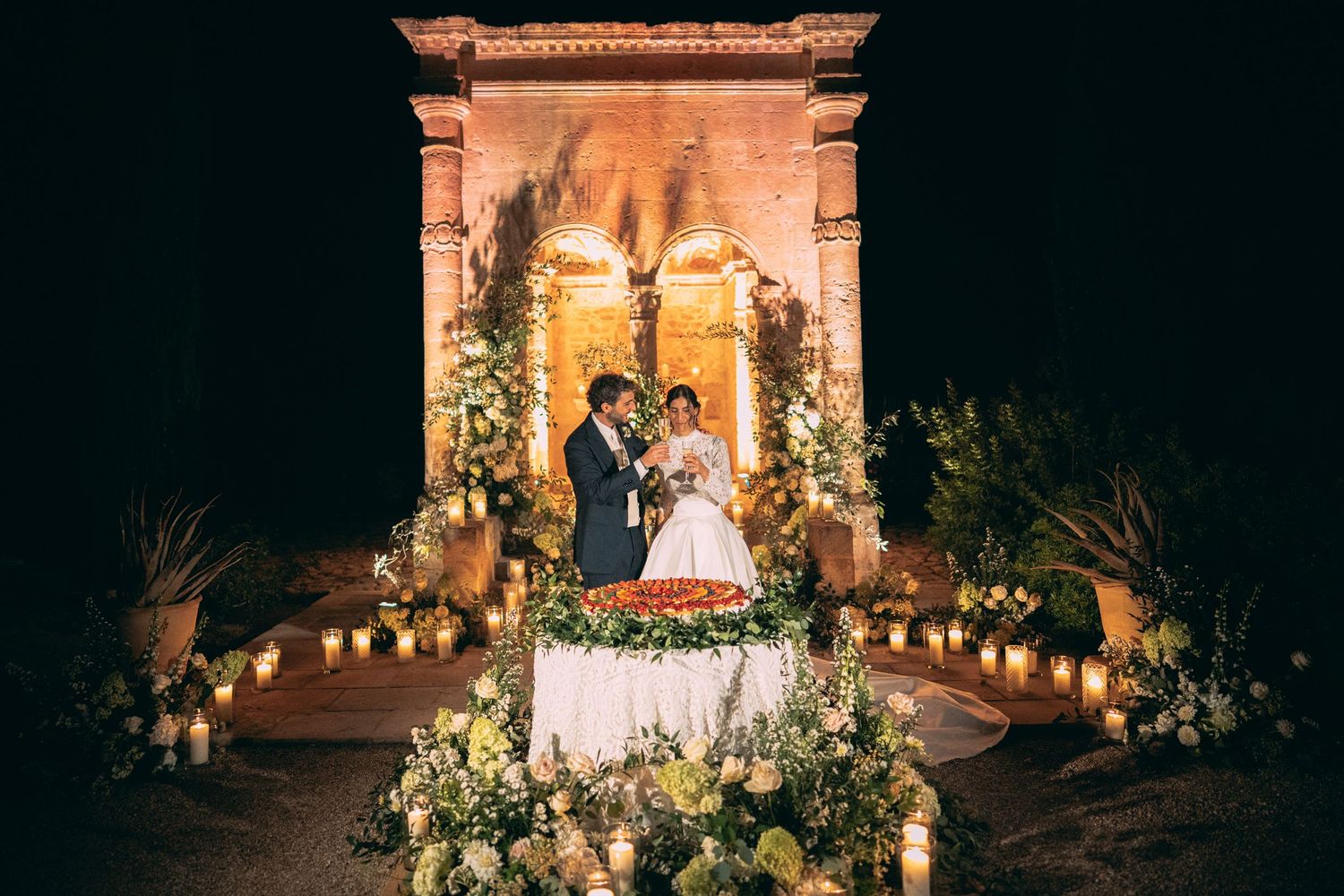Romantic outdoor wedding reception at night with couple at floral table, candlelit arch backdrop and lush greenery.