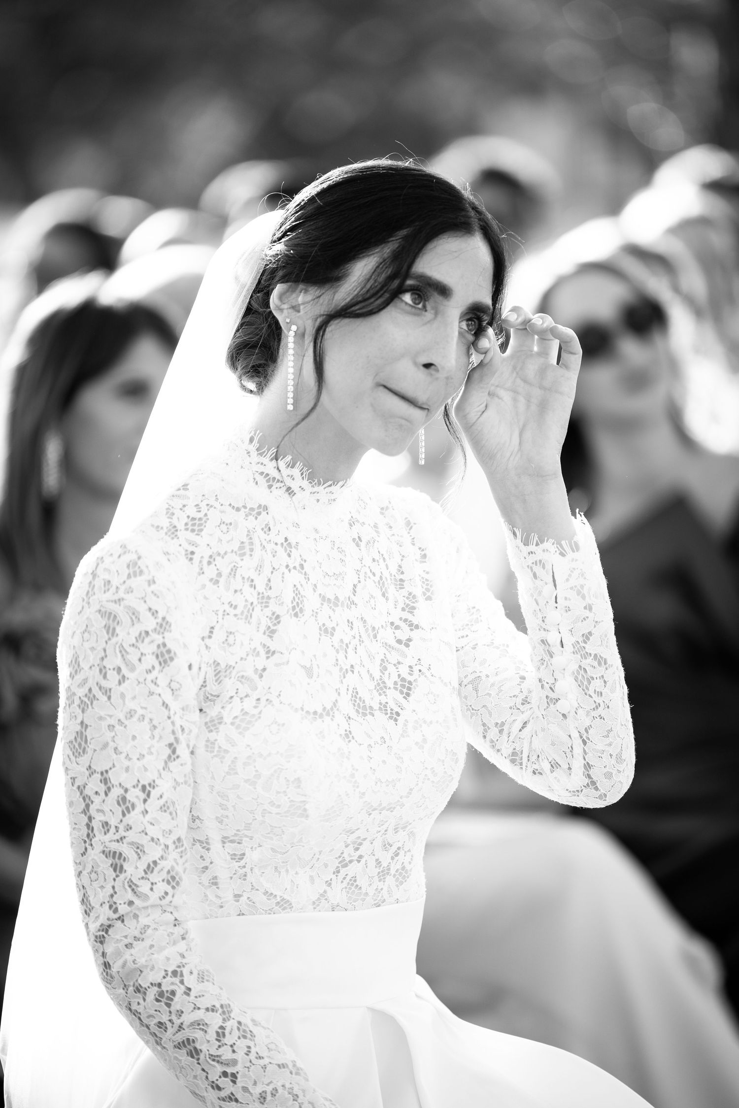 Black and white photo of an emotional bride in lace gown wiping tears during wedding ceremony.