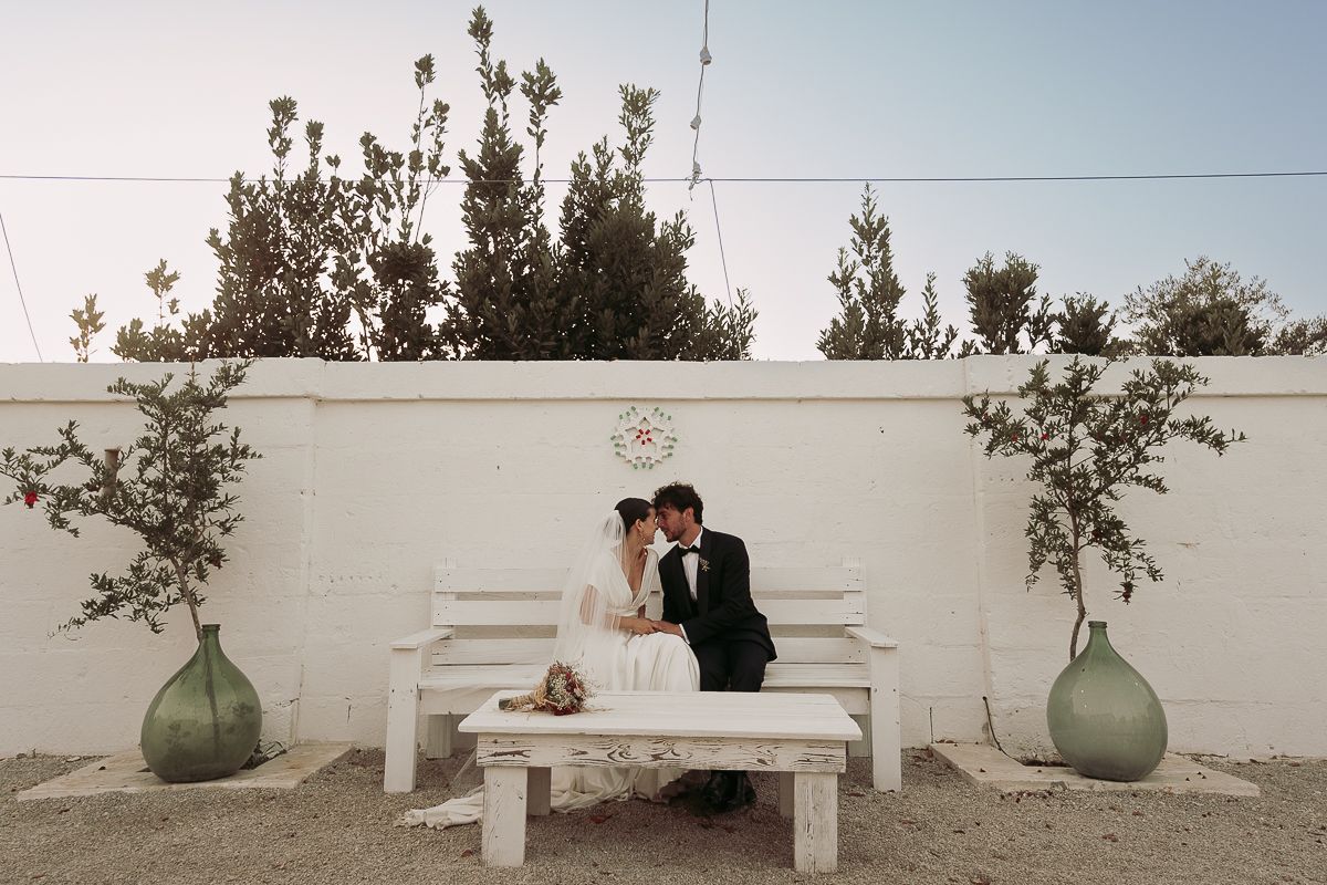 Bride and groom share a kiss on a white bench surrounded by lush greenery and large vases at an outdoor wedding venue.