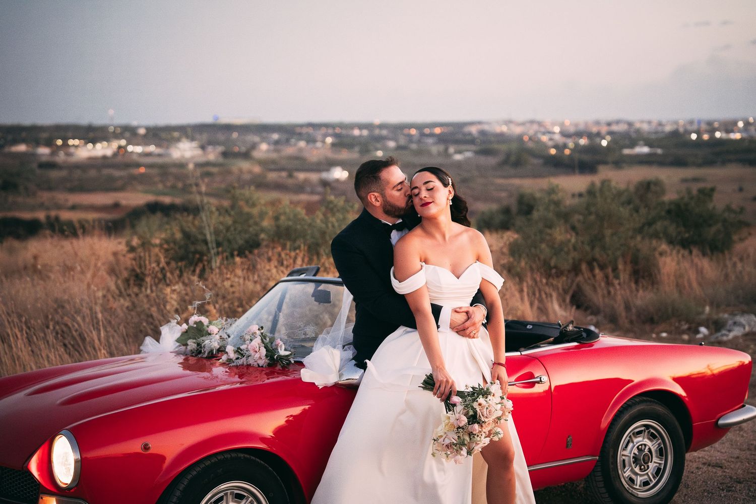 Bride and groom embracing beside a classic red convertible sports car at sunset in a romantic outdoor wedding photo.