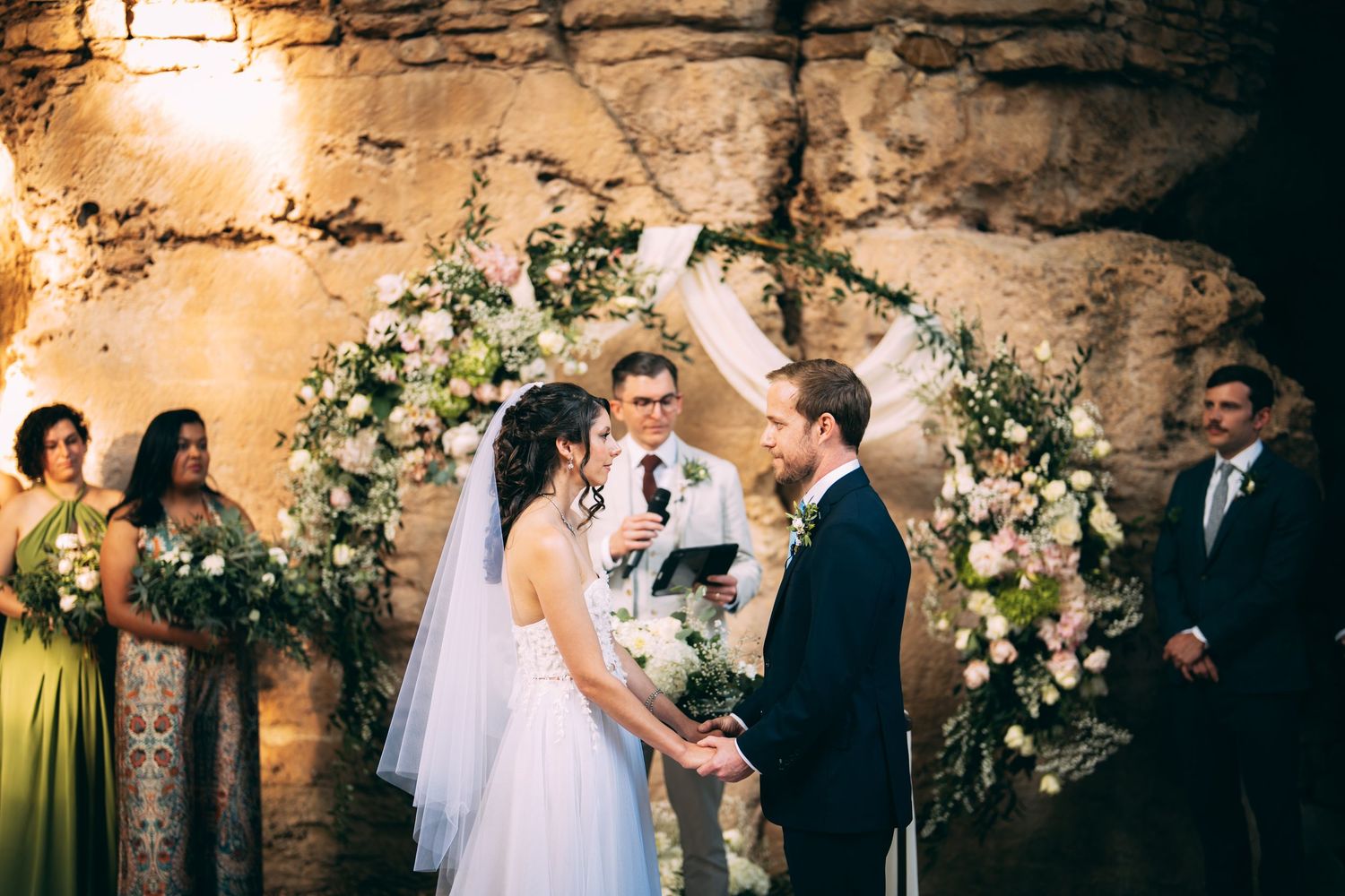 Bride and groom exchange vows at a floral arch ceremony in a rustic stone venue with bridesmaids nearby.