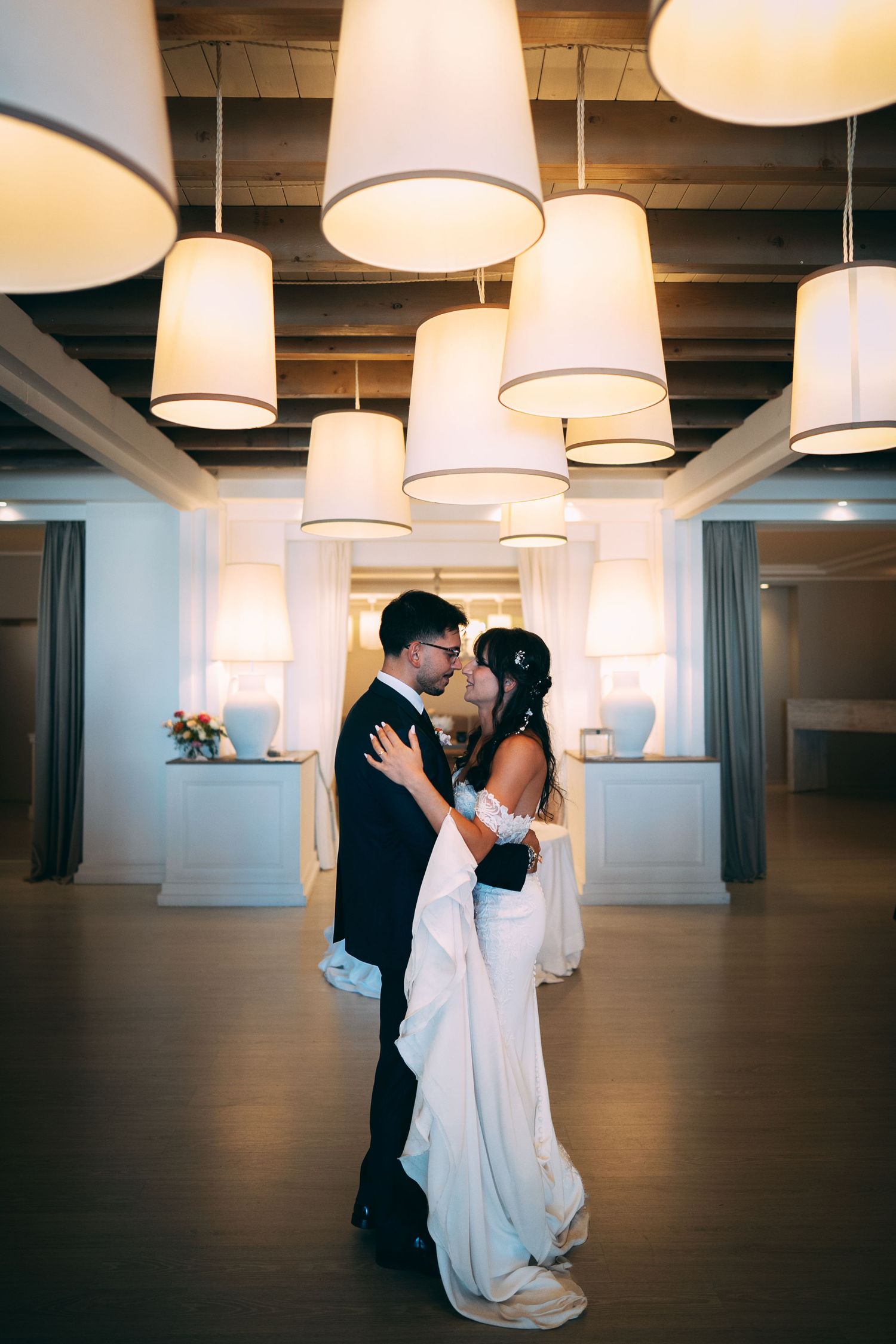 Bride and groom share a romantic kiss under elegant hanging lamp shades in a softly lit wedding reception venue.