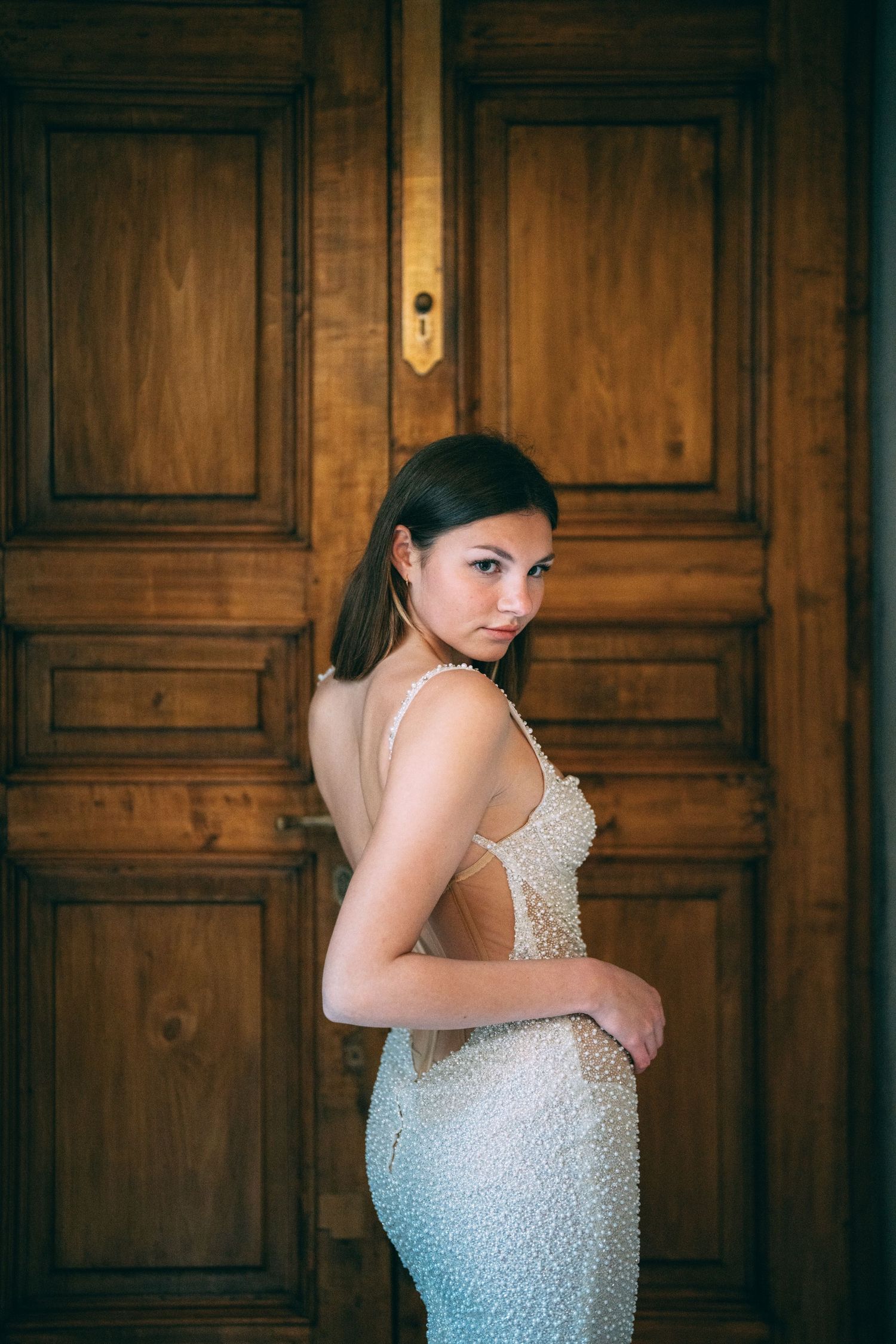 Young woman in elegant backless pearl gown poses gracefully before ornate wooden doors.