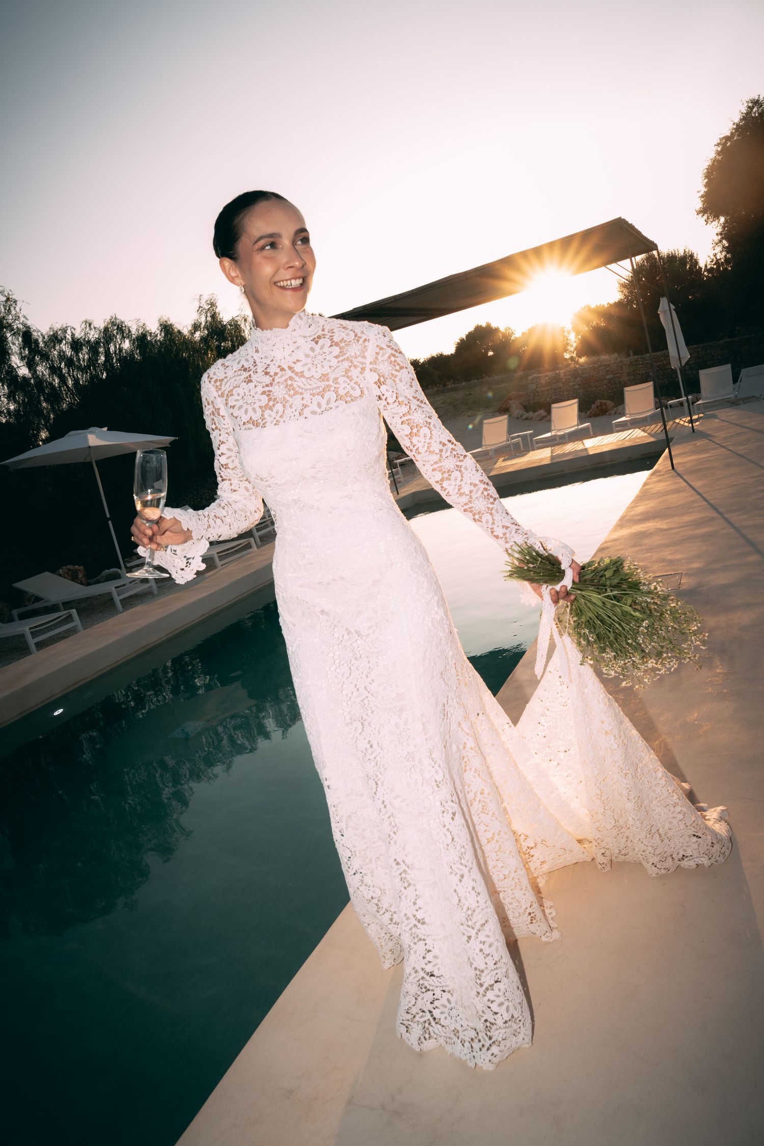 Bride in white lace long-sleeve gown holding bouquet and champagne glass by pool at sunset.