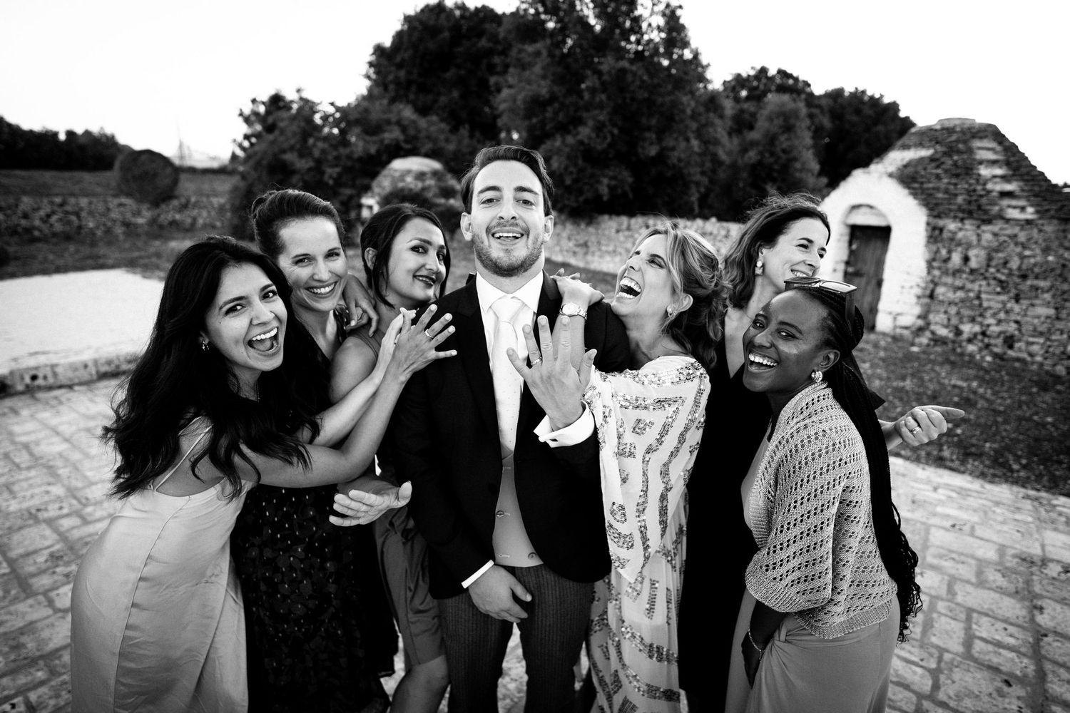 Black and white photo of a groom surrounded by laughing bridesmaids at an outdoor wedding venue.