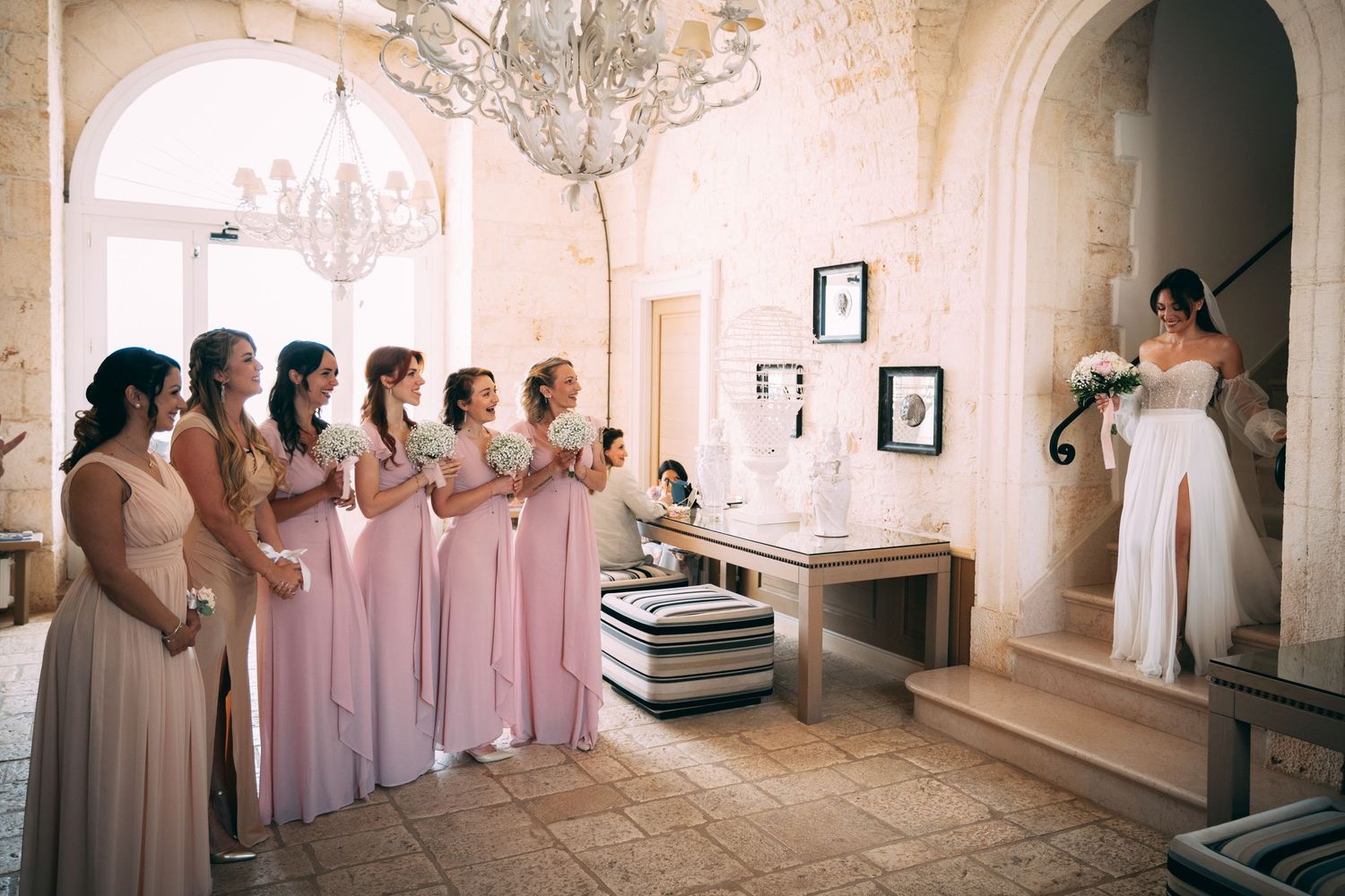 Bridesmaids in pink gowns stand beside a bride in white inside an elegant venue with chandelier.