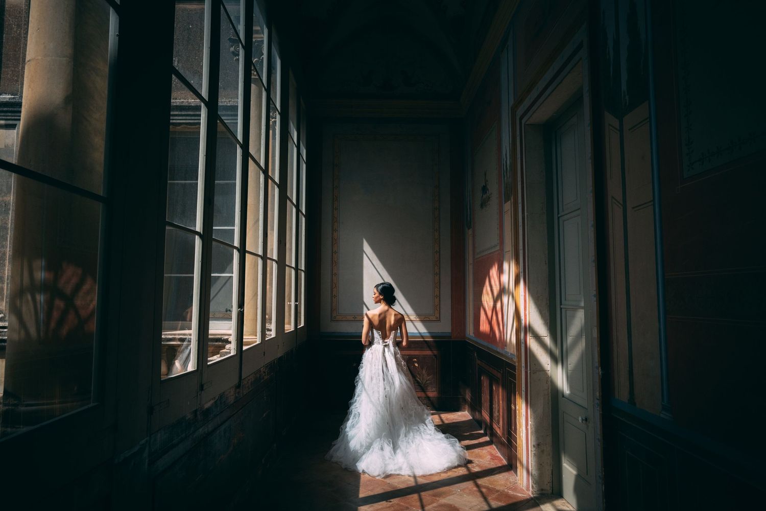 Sunlight streams through tall windows onto a person in a white wedding dress standing in an elegant corridor.