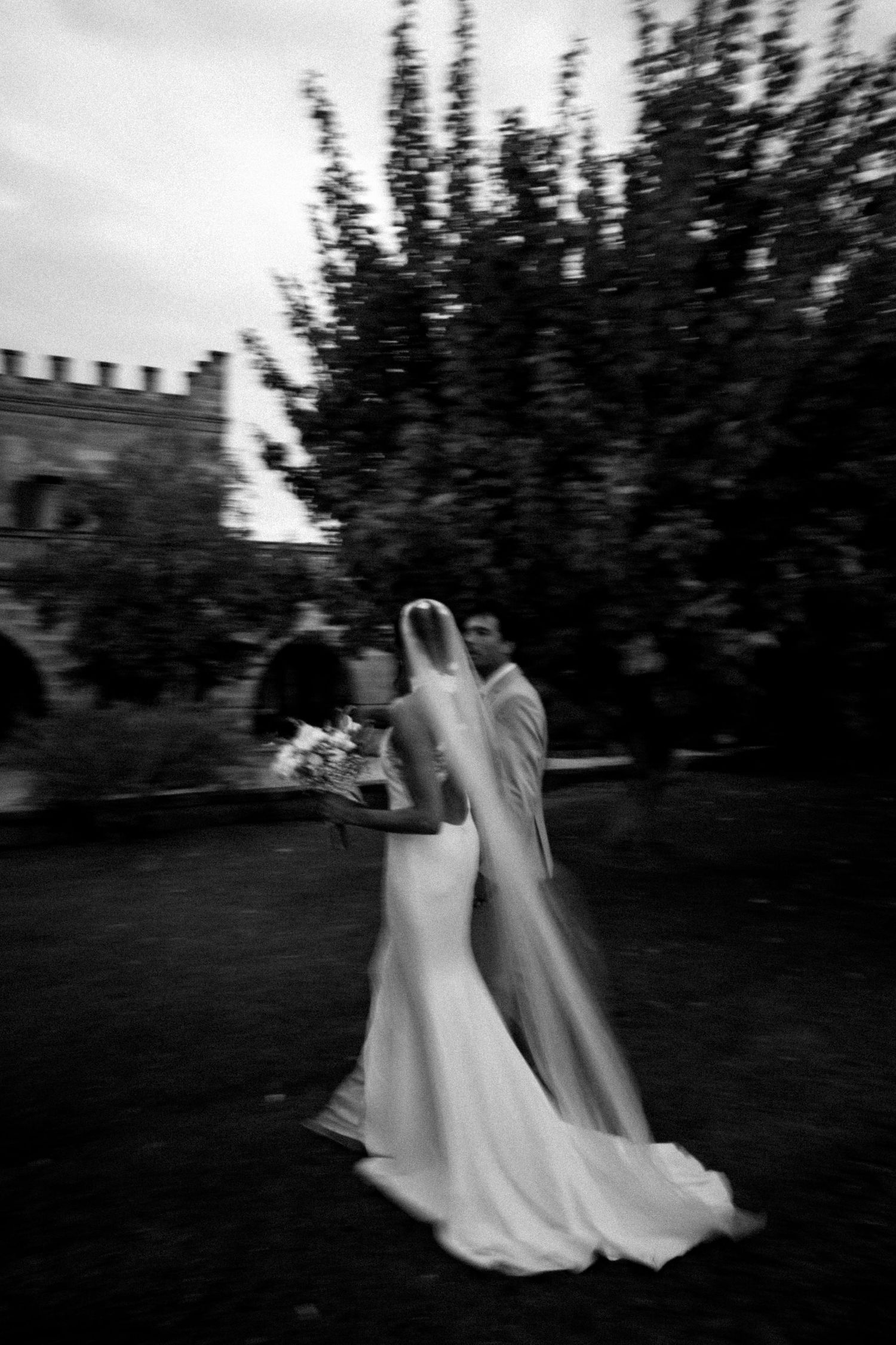 Two figures in flowing white dresses walk together through a garden in a haunting black and white photograph.