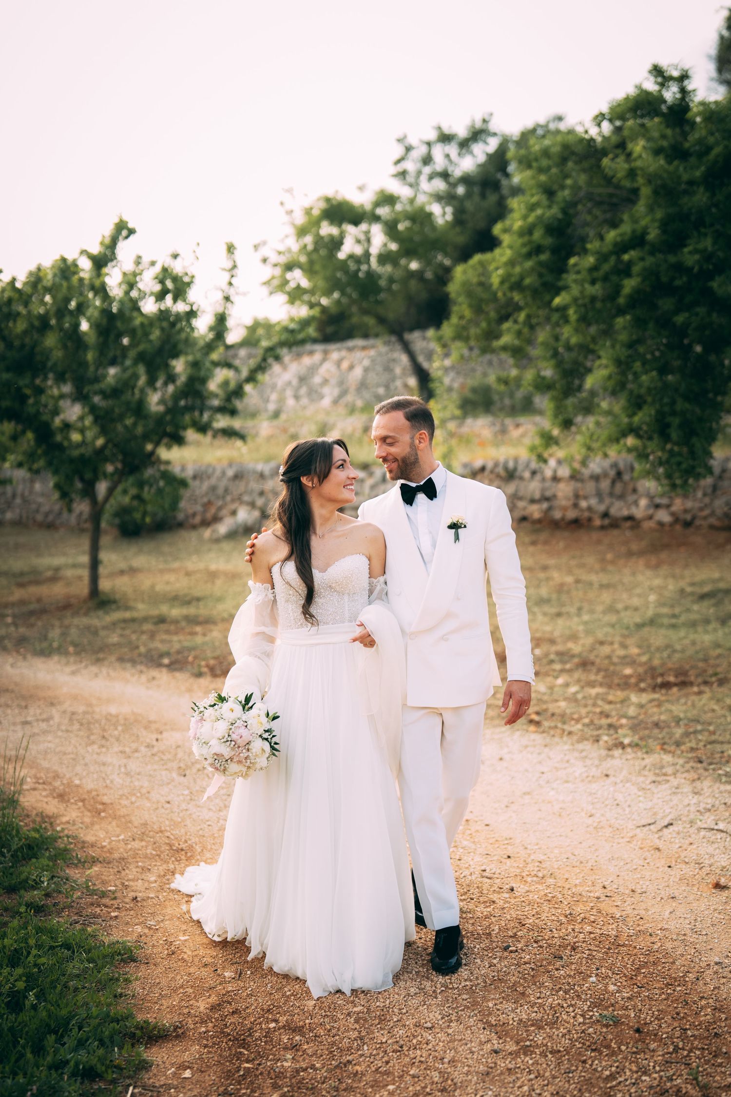 Bride and groom walk hand in hand on a sunlit gravel path, surrounded by lush green trees on their wedding day.
