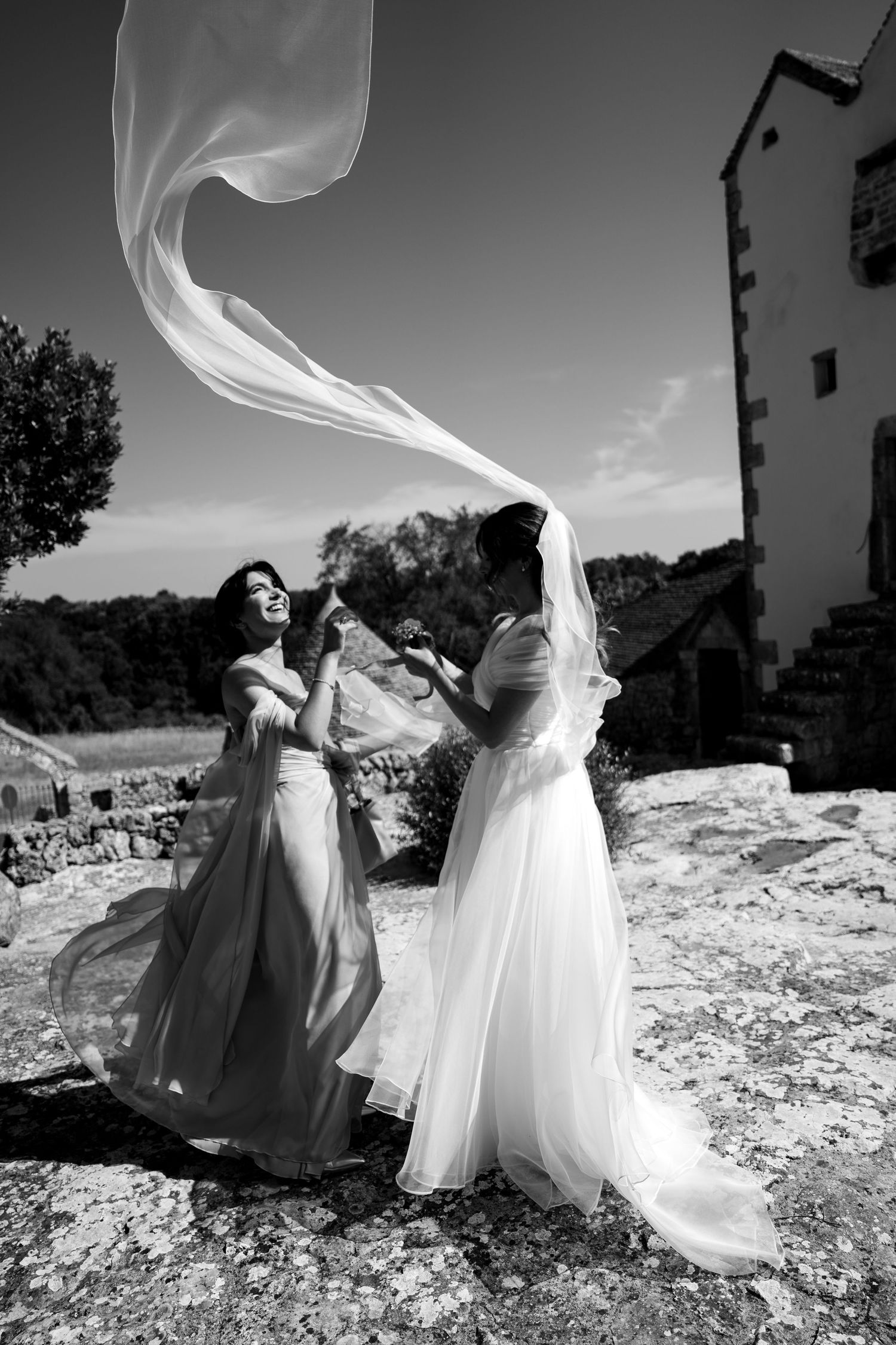 Black and white photo of bride and bridesmaid laughing as wedding veil blows in wind near stone building.