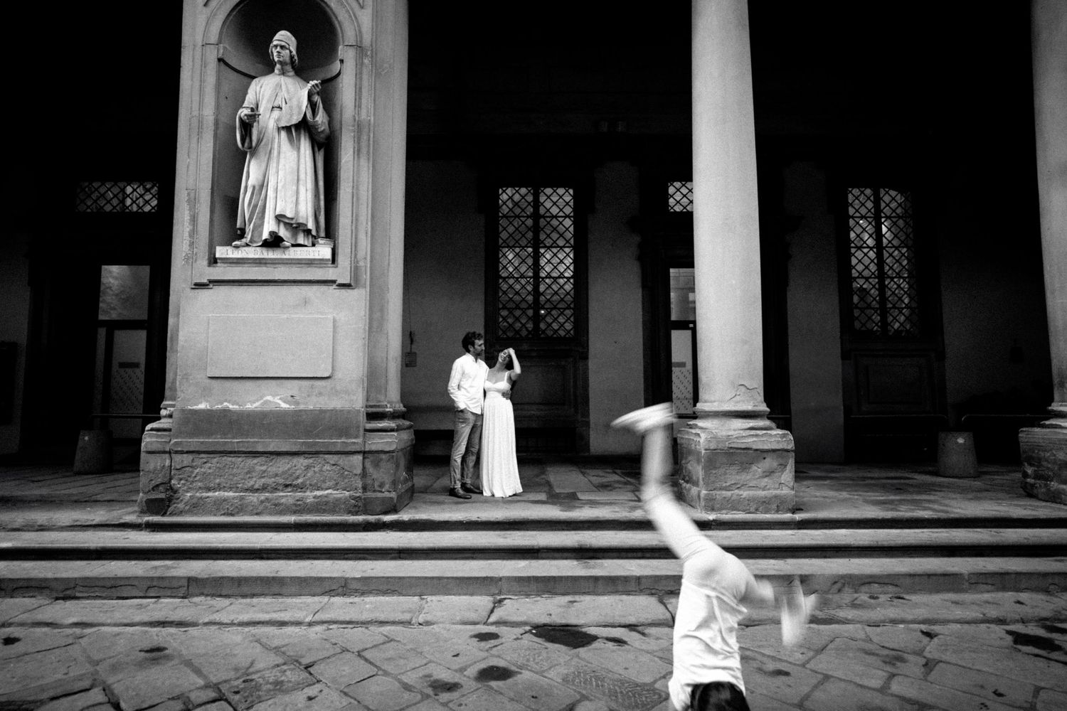 Black and white photo of a couple embracing near a statue while a person does a cartwheel in the foreground.